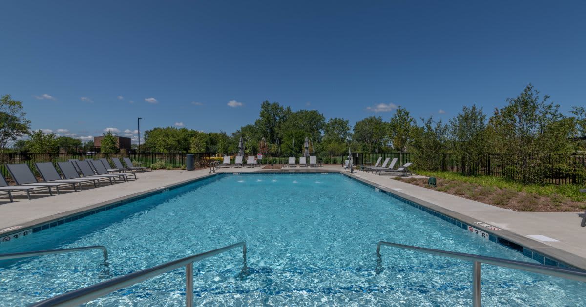 Outdoor swimming pool with clear blue water, metal railings, lounge chairs, and trees under blue sky.