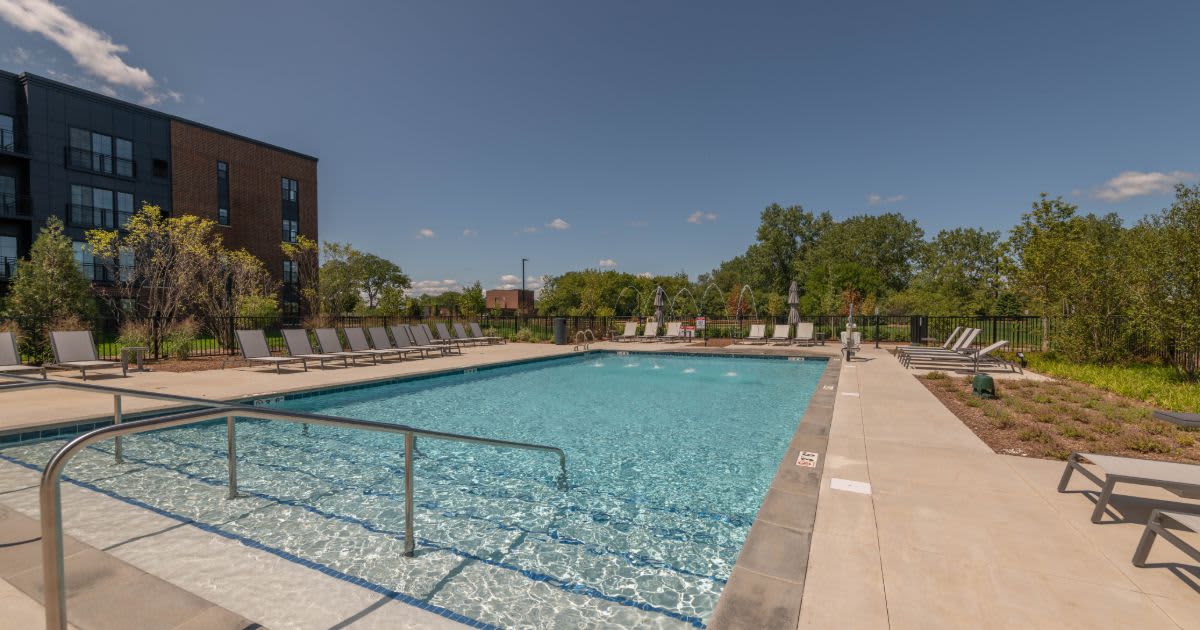 Outdoor swimming pool with metal safety railing, concrete deck, lounge chairs, and modern brick apartment building under blue sky.