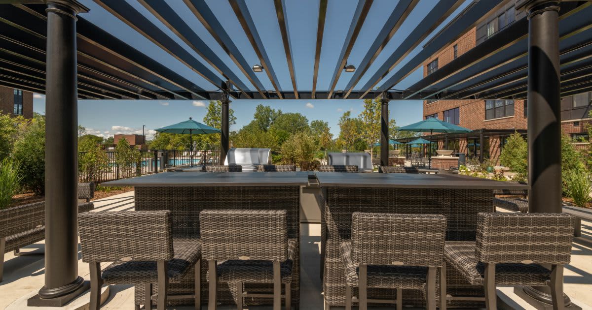 Rooftop terrace with woven chairs, dining tables under a black pergola, brick buildings and green landscaping visible in background.