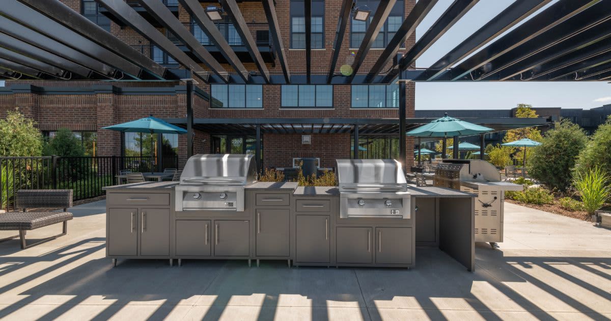 Rooftop terrace with two stainless steel gas grills, gray cabinetry, pergola overhead, and turquoise umbrellas overlooking brick building and landscaped grounds.