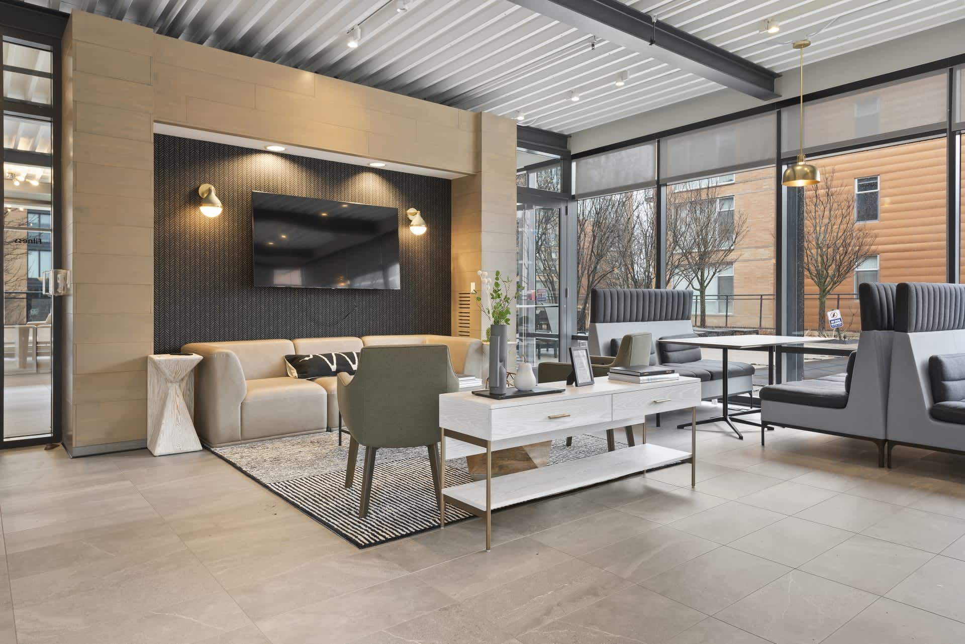 Modern apartment lounge with beige sofa, green accent chair, white coffee table, black accent wall with TV, and large windows overlooking trees.
