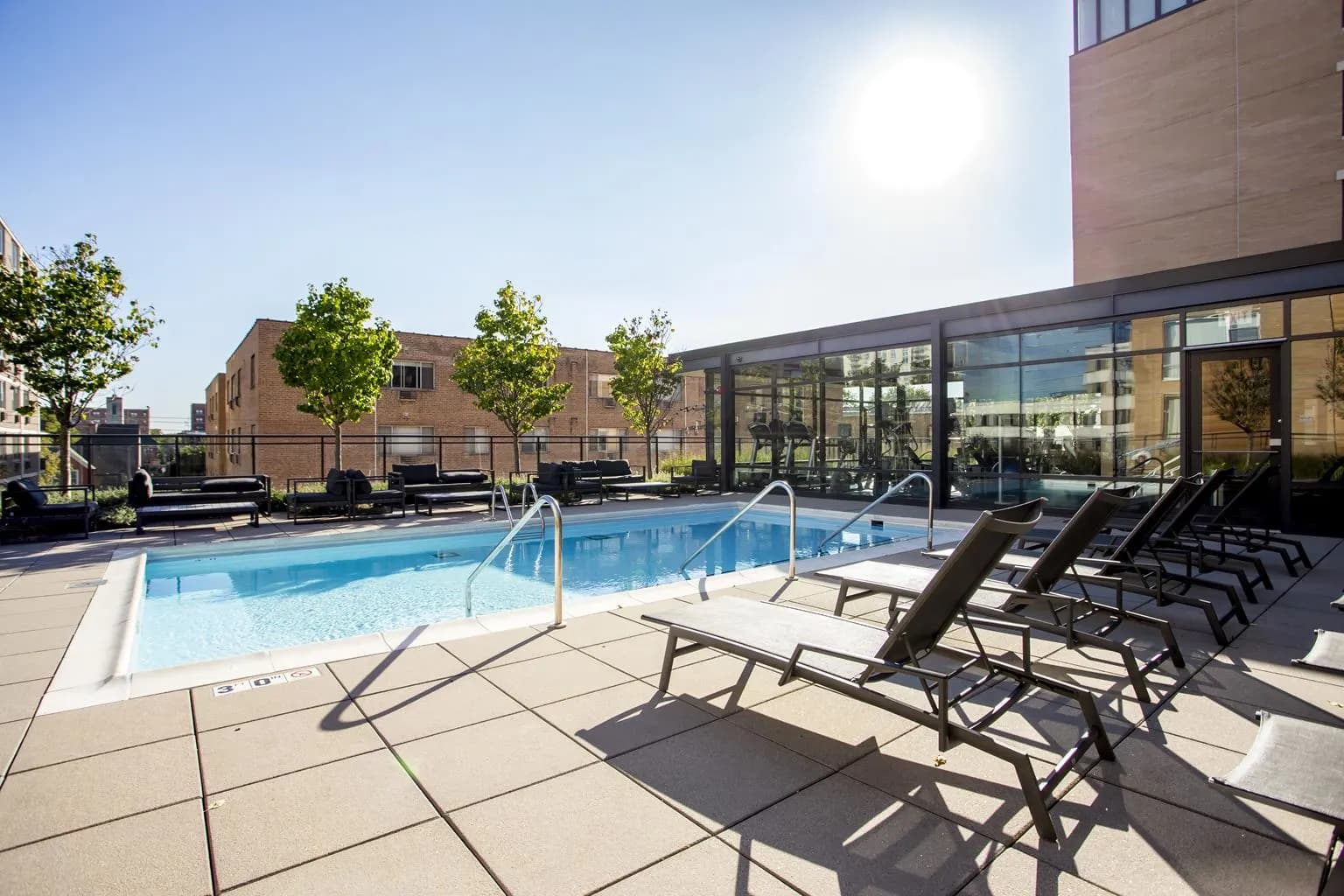Rooftop pool with metal lounge chairs, white railings, and modern brick building with glass windows under clear sky.