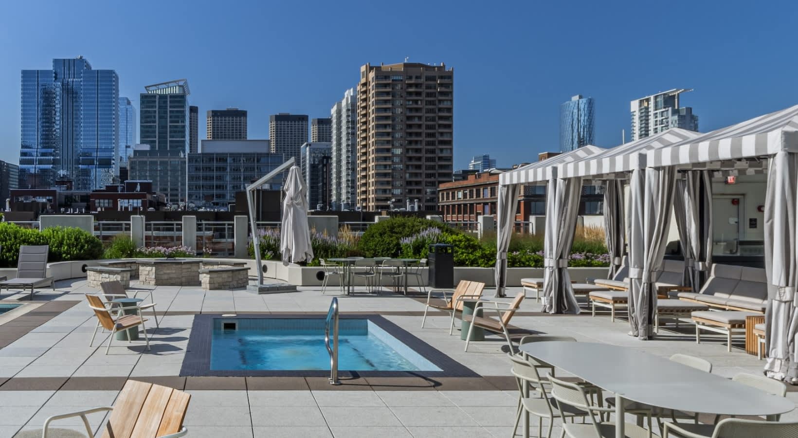 Rooftop pool deck with rectangular lap pool, wooden lounge chairs, white cabanas with curtains, and Chicago skyline in background.