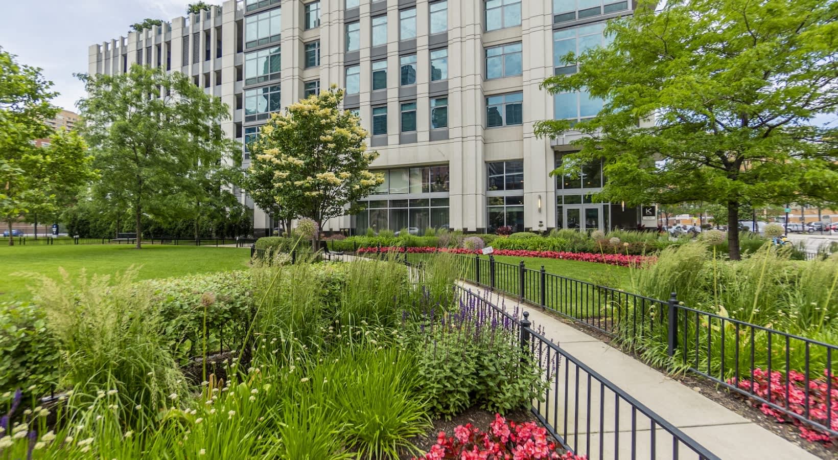 Modern residential building with manicured lawn, ornamental grasses, red flowers, black metal railing, and mature trees.
