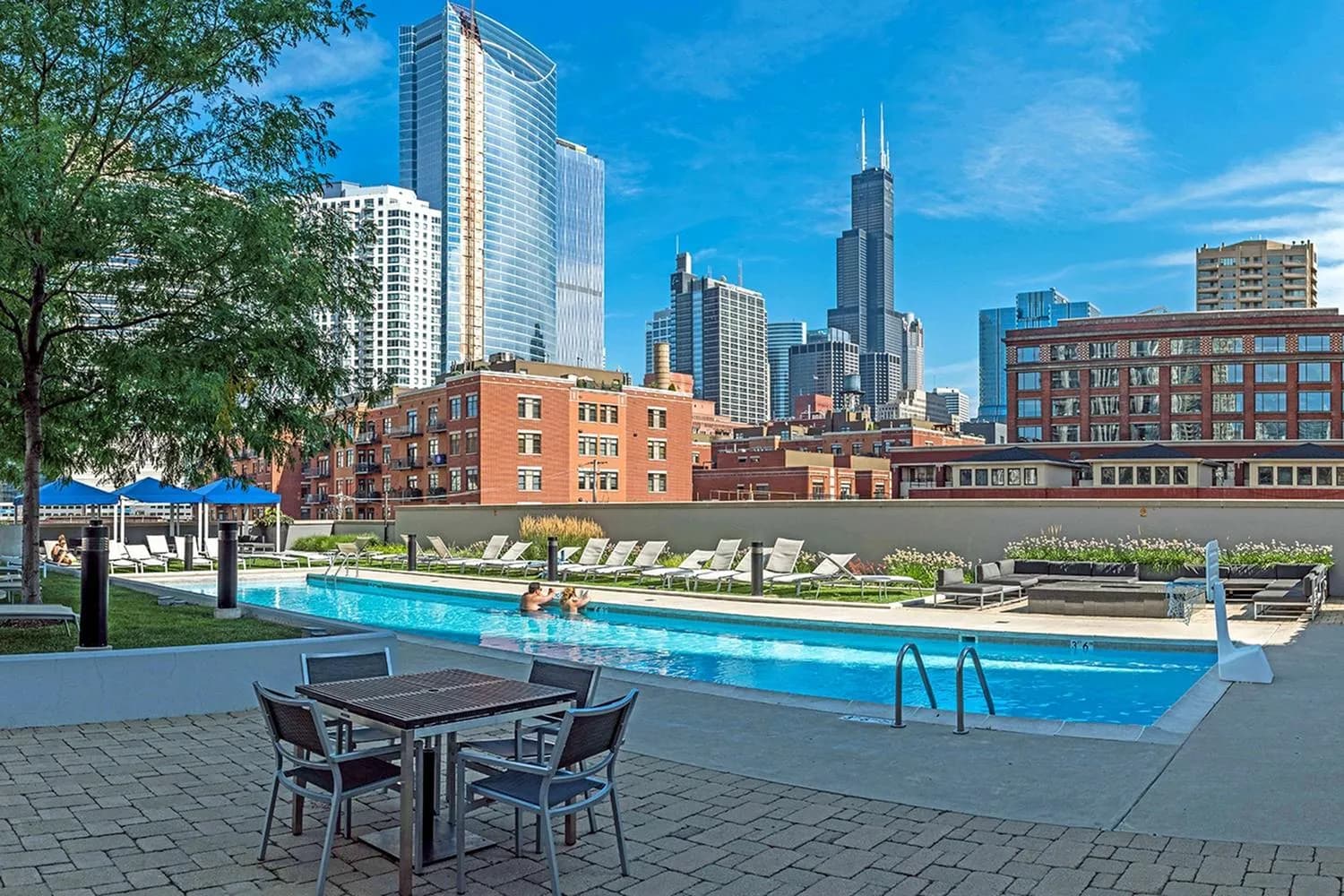 Rooftop pool with blue water, lounge chairs, dining table, and Chicago skyline featuring Willis Tower in background.