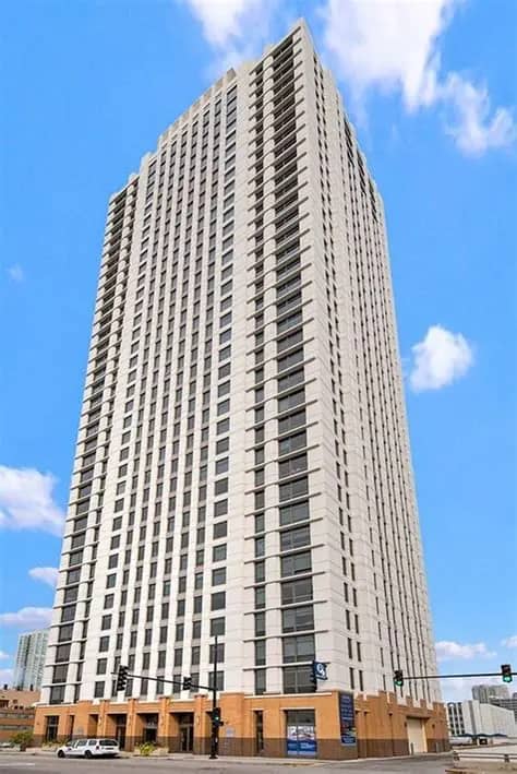 Tall residential tower with regular window pattern, tan and white facade, ground-floor retail space, blue sky background.