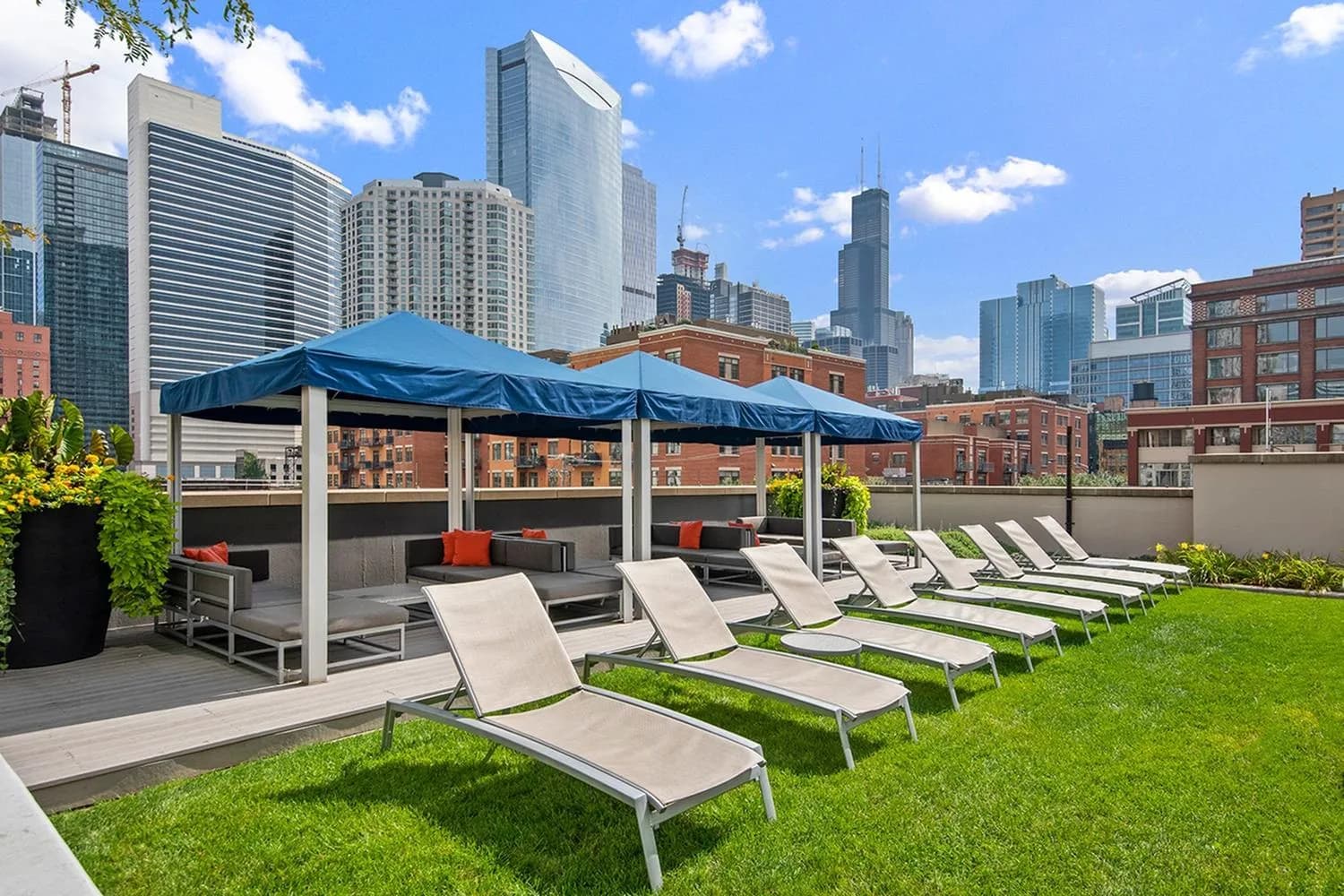 Rooftop terrace with rows of white lounge chairs on grass, blue canopy structure with seating, and Chicago skyline in background.