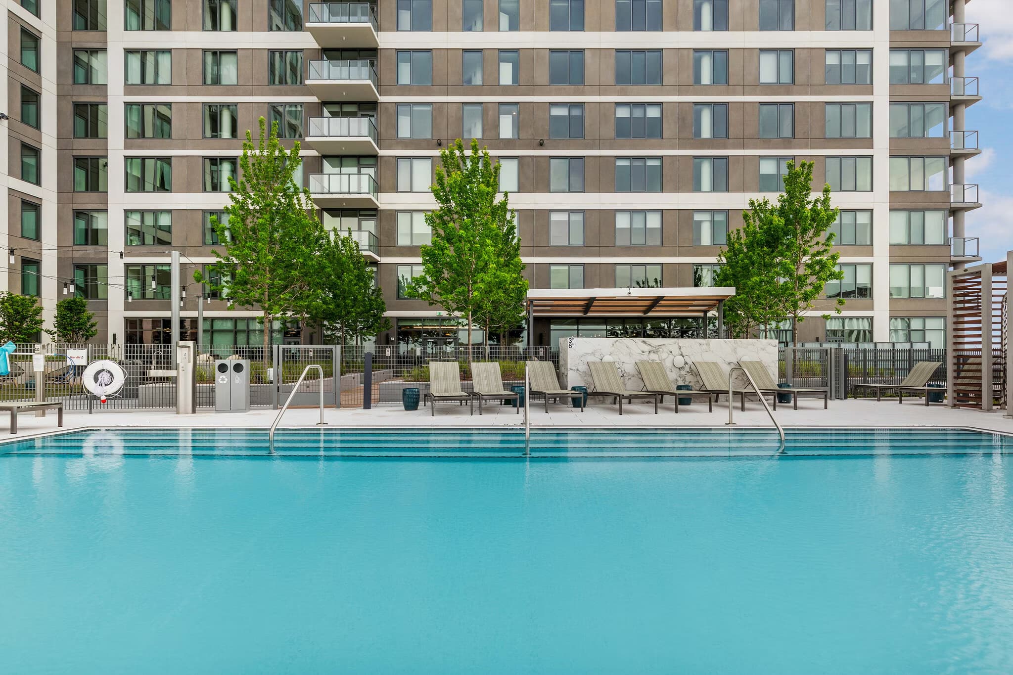 Outdoor swimming pool with clear turquoise water, lounge chairs, and tall green trees in front of a modern multi-story residential building.