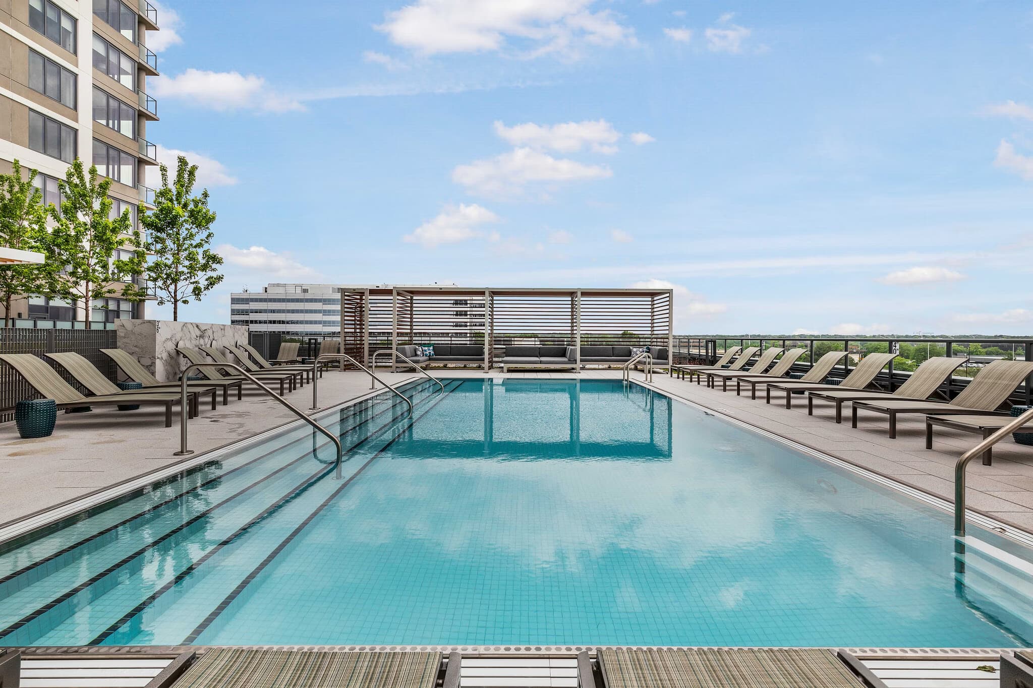 Outdoor swimming pool with lane markers, surrounded by lounge chairs and a modern residential building under blue sky.