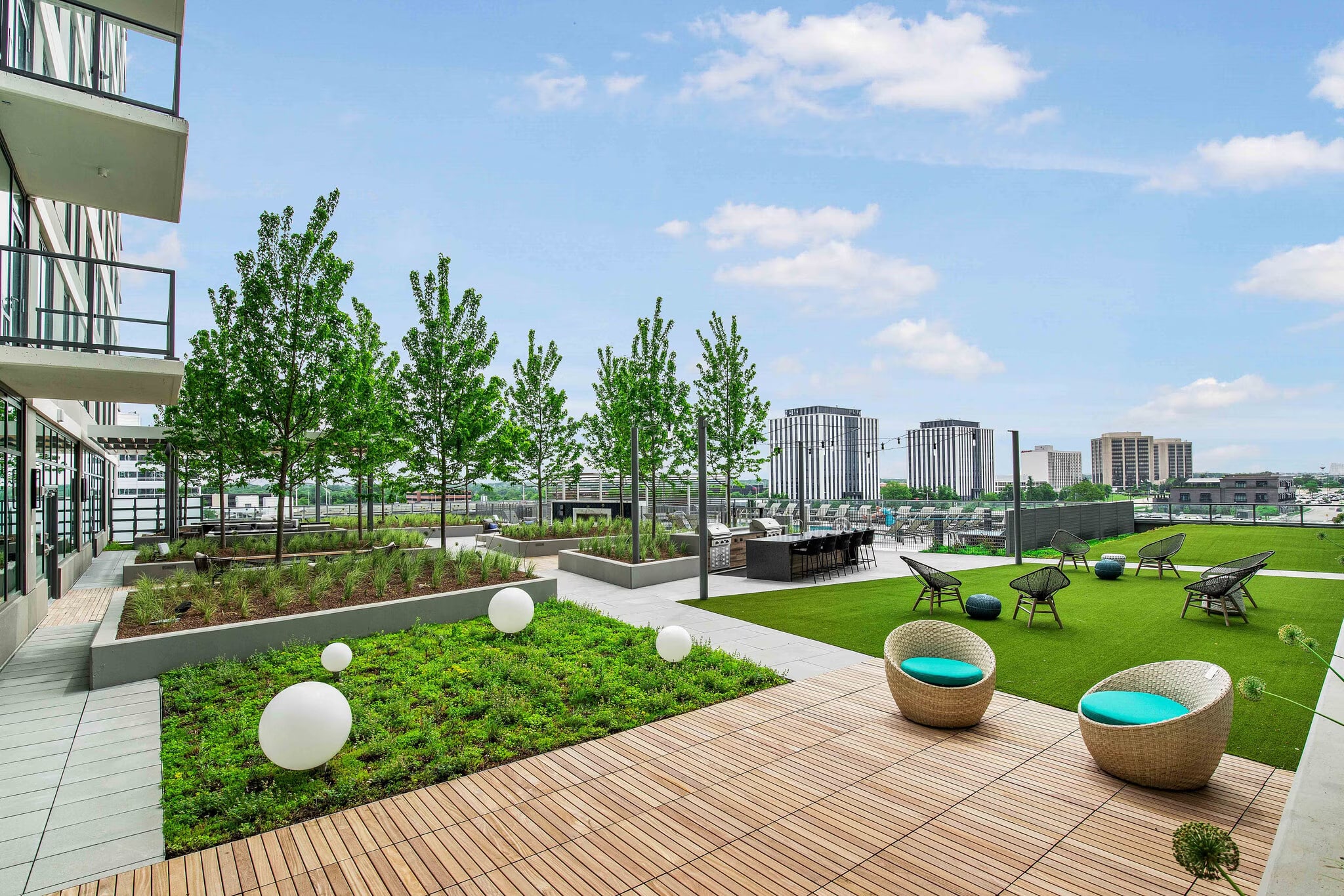 Rooftop terrace with green lawn, woven lounge chairs with turquoise cushions, white decorative spheres, young trees, and city skyline views.