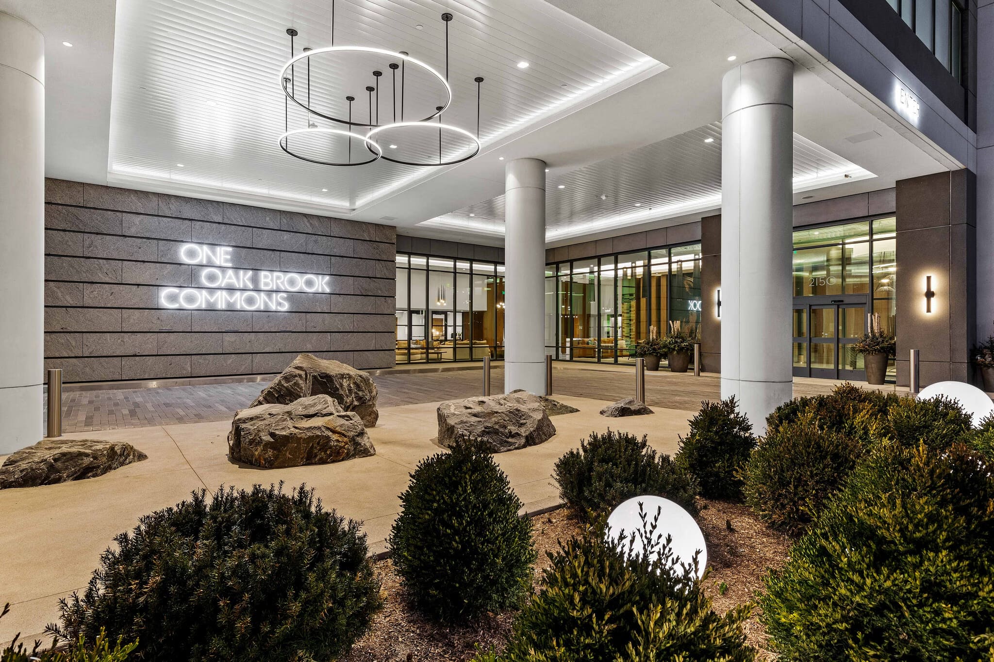 Modern building lobby with One Oak Brook Commons neon sign, white columns, decorative boulders, and manicured shrubs under recessed lighting.