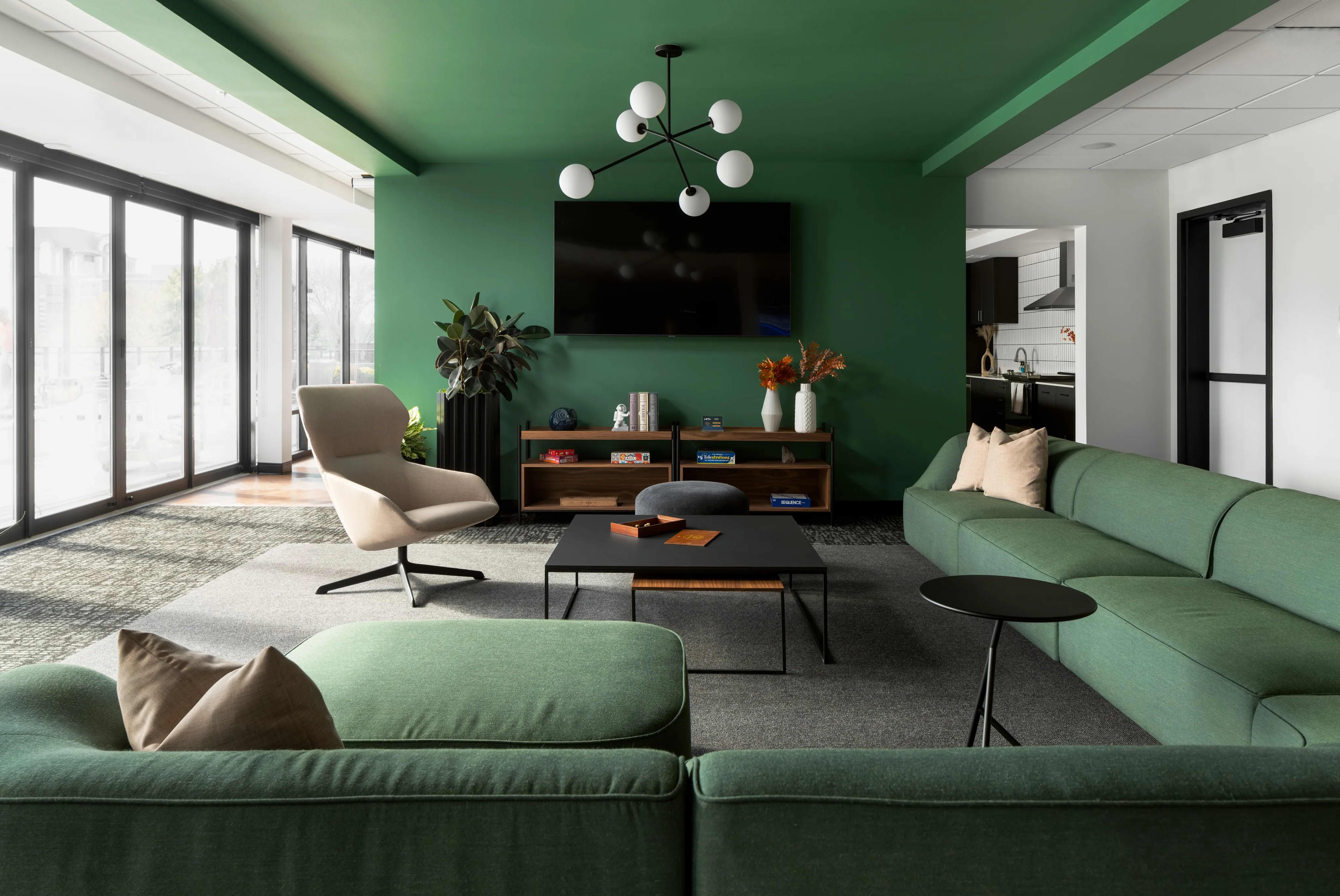 Living room with sage green walls and ceiling, green sectional sofa, beige accent chair, black coffee table, wooden shelving unit, and glass doors opening to outdoor space.