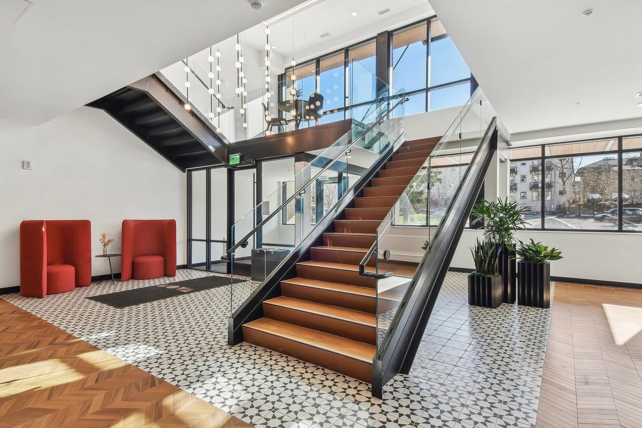 Modern lobby with wooden staircase, glass railings, red seating pods, geometric floor tiles, and floor-to-ceiling windows overlooking the street.