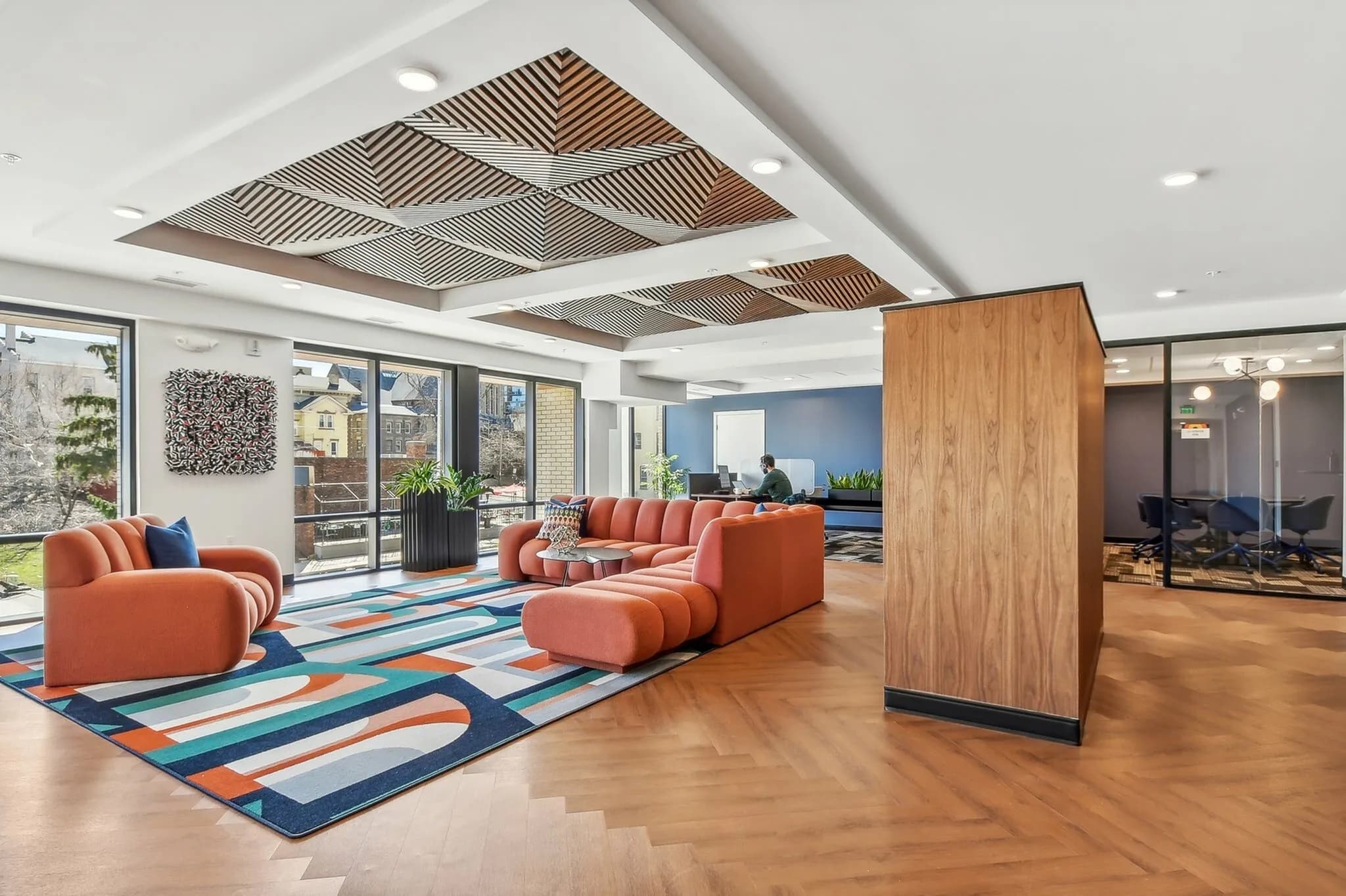 Open lounge area with curved orange seating, geometric wood ceiling panels, colorful area rug, and floor-to-ceiling windows overlooking the city.
