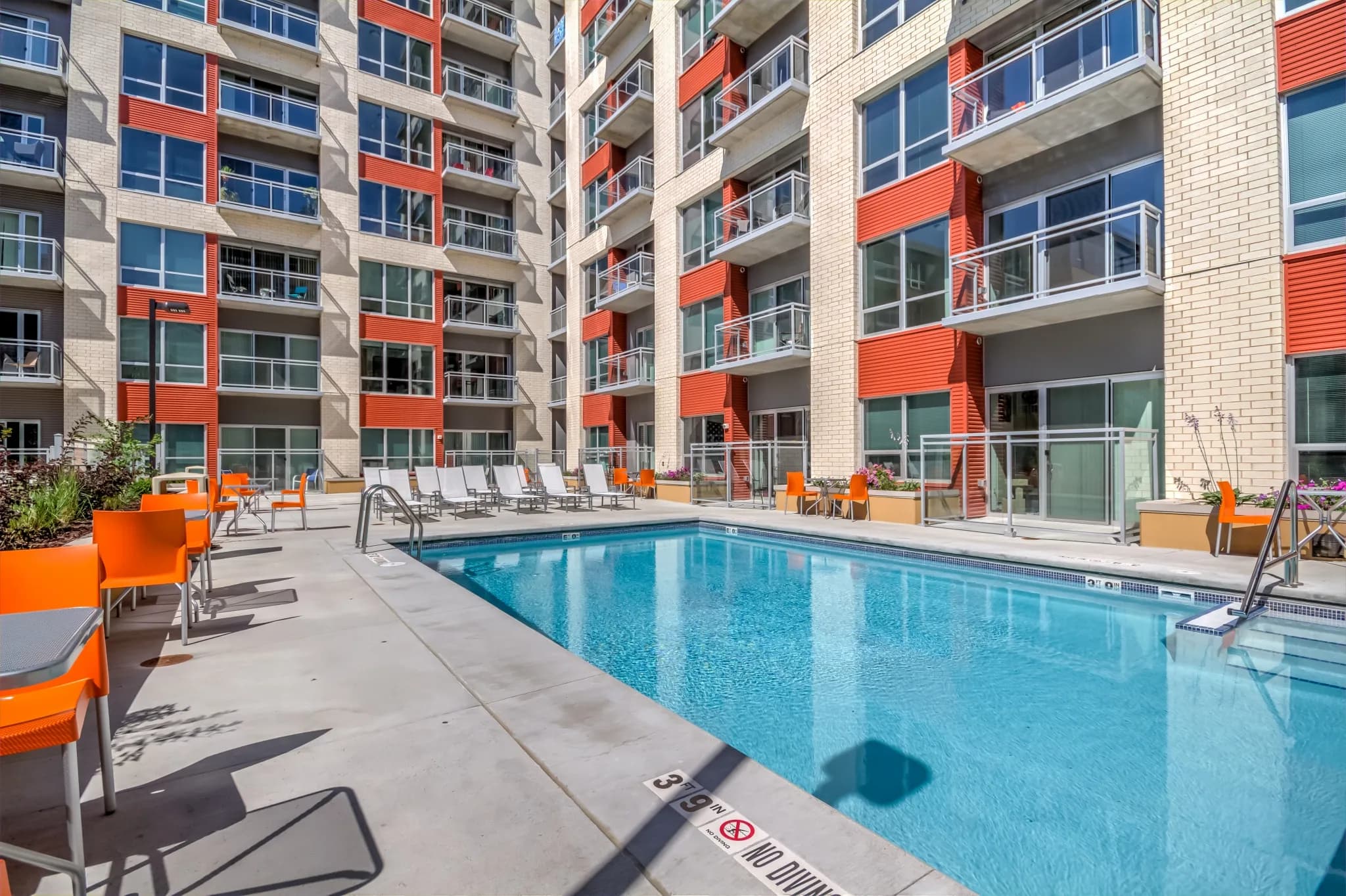Outdoor swimming pool with bright blue water, surrounded by concrete deck, orange lounge chairs, and multi-story residential building with red and cream facade.