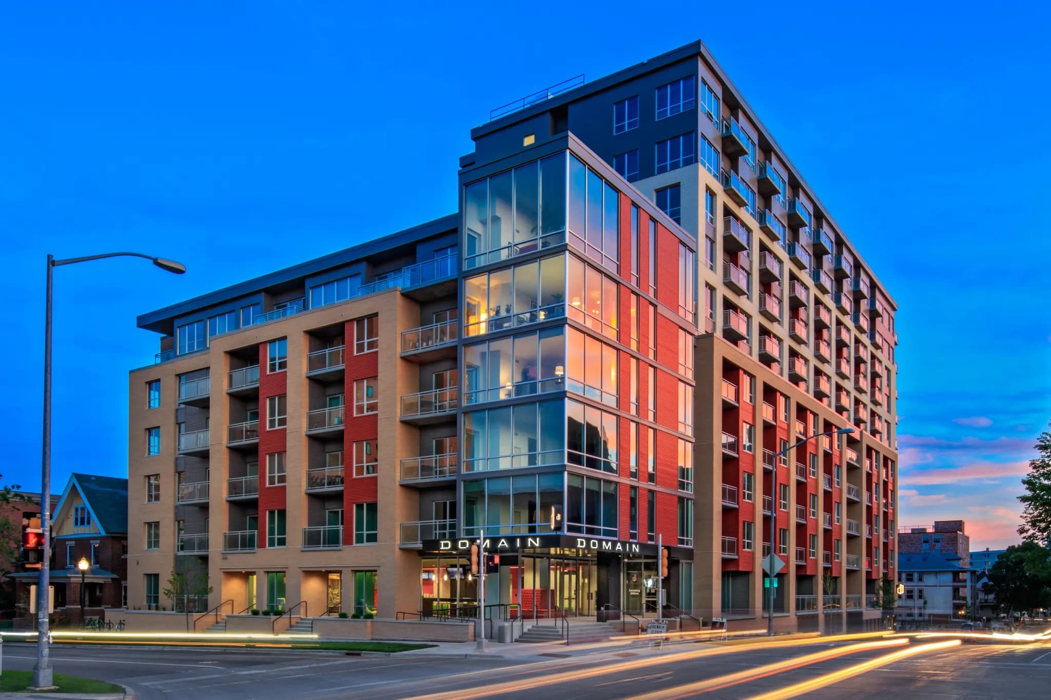 Modern mixed-use building with red and tan facade, ground-floor retail, and residential units above at dusk.