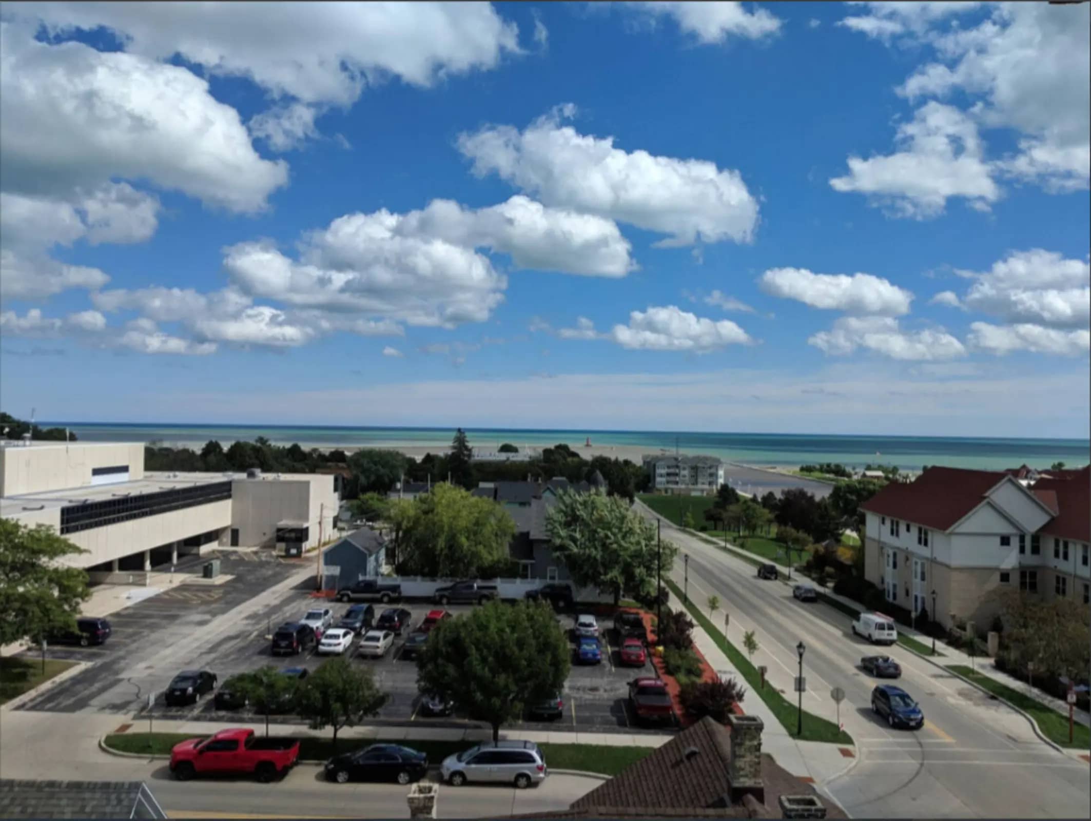 Elevated view of parking lot and street with Lake Michigan and sandy beach visible in the distance under blue sky with white clouds.
