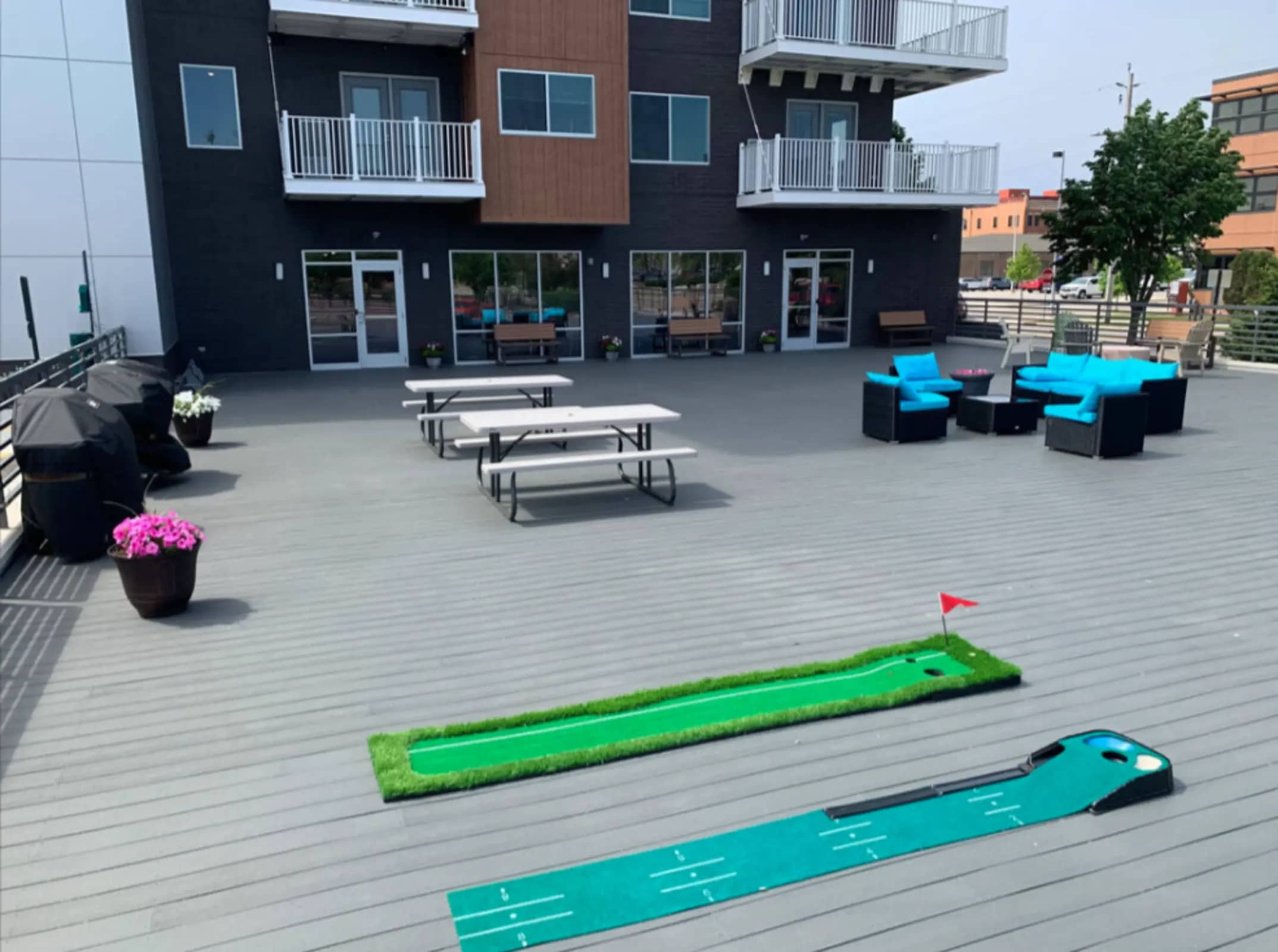 Rooftop deck with mini golf course, turquoise lounge seating, picnic tables, and modern apartment building with black and wood facade.