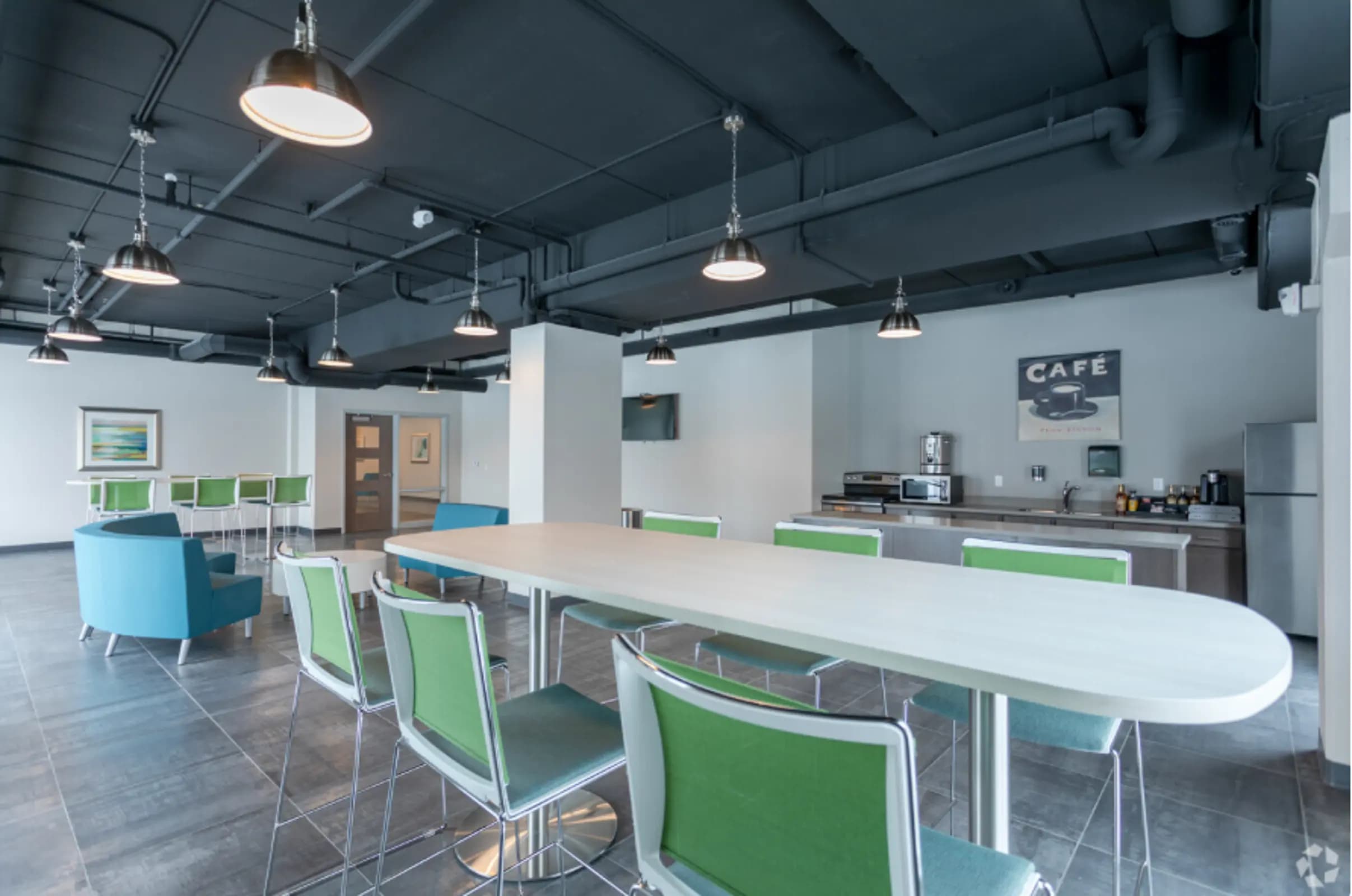 Open-concept common kitchen and dining area with white countertops, green and blue chairs, stainless steel appliances, and dark ceiling with pendant lights.