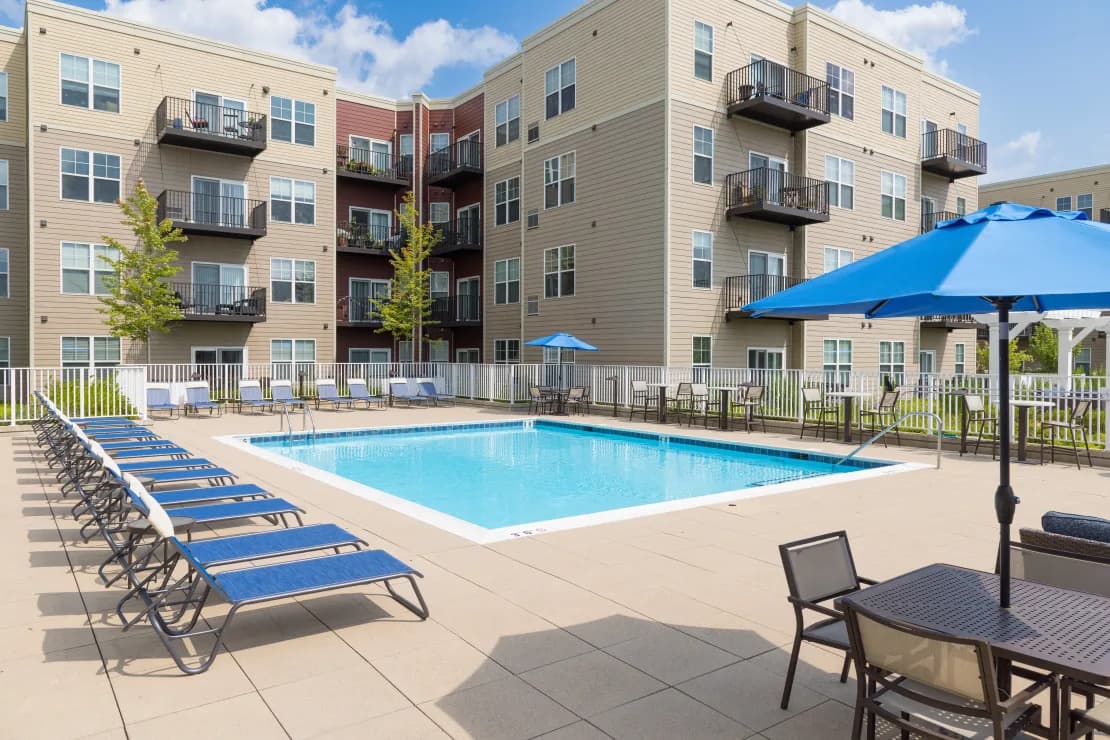 Outdoor swimming pool with blue lounge chairs, umbrellas, and dining tables, backed by multi-story residential buildings under clear sky.