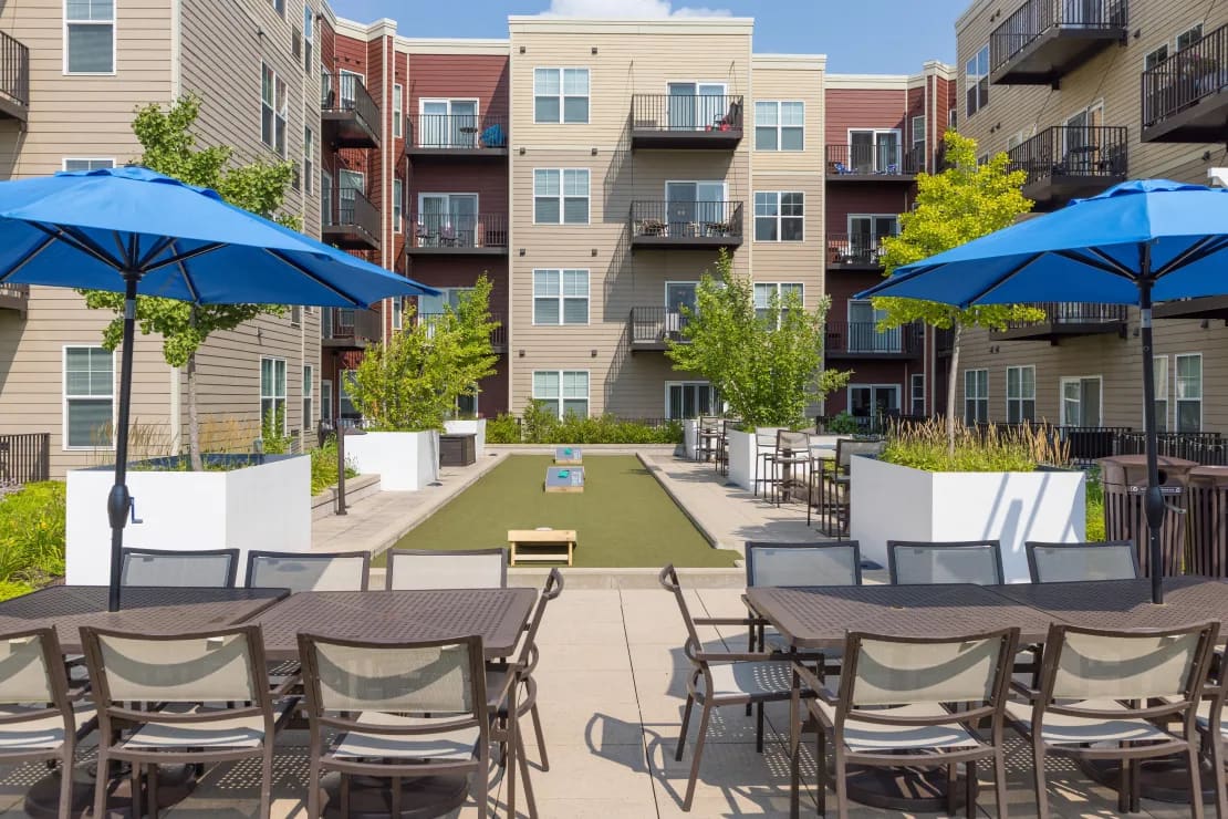 Outdoor courtyard with bocce ball court, lounge chairs, blue umbrellas, and apartment buildings in background.