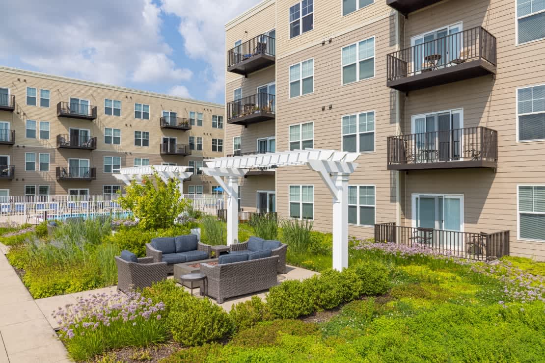 Outdoor courtyard with wicker seating, white pergola, and landscaped gardens with purple flowers and green shrubs, flanked by tan apartment buildings.