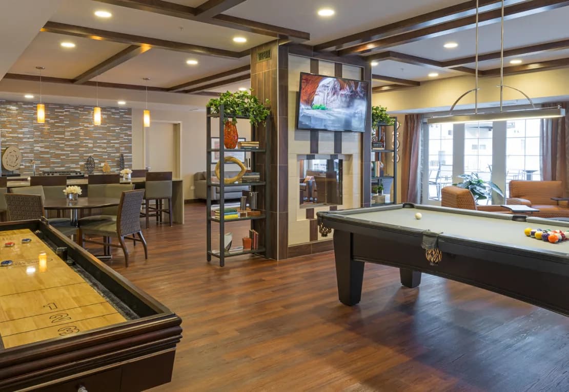 Recreation room with pool table, shuffleboard, wooden flooring, and lounge seating areas with decorative shelving and wall-mounted TV.