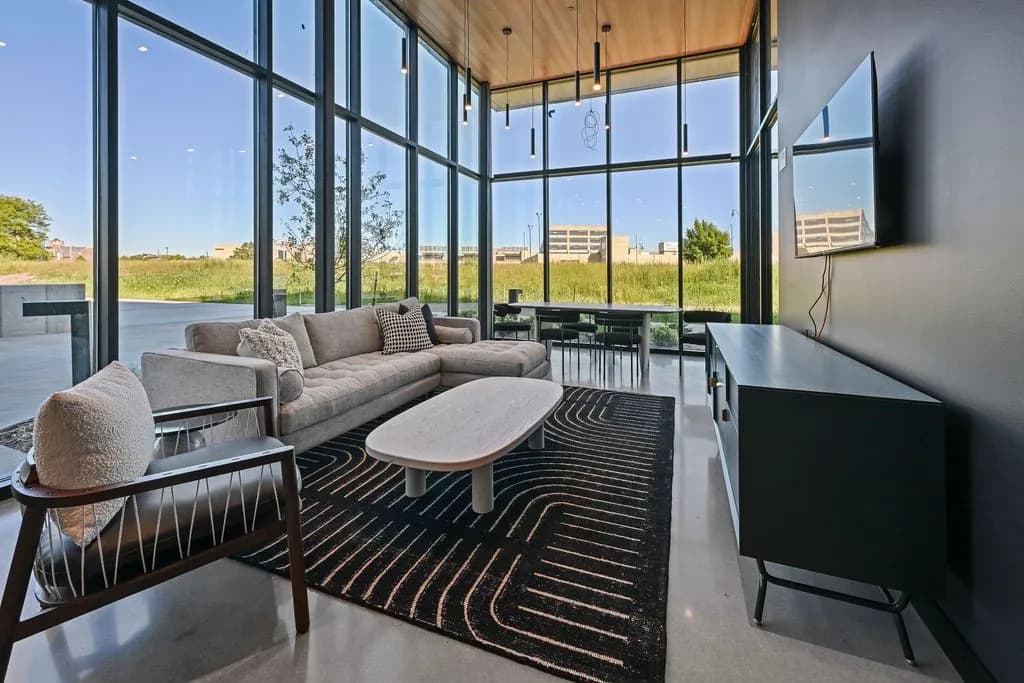 Living room with gray sectional sofa, wooden coffee table, geometric patterned rug, and expansive black-framed windows with city views.