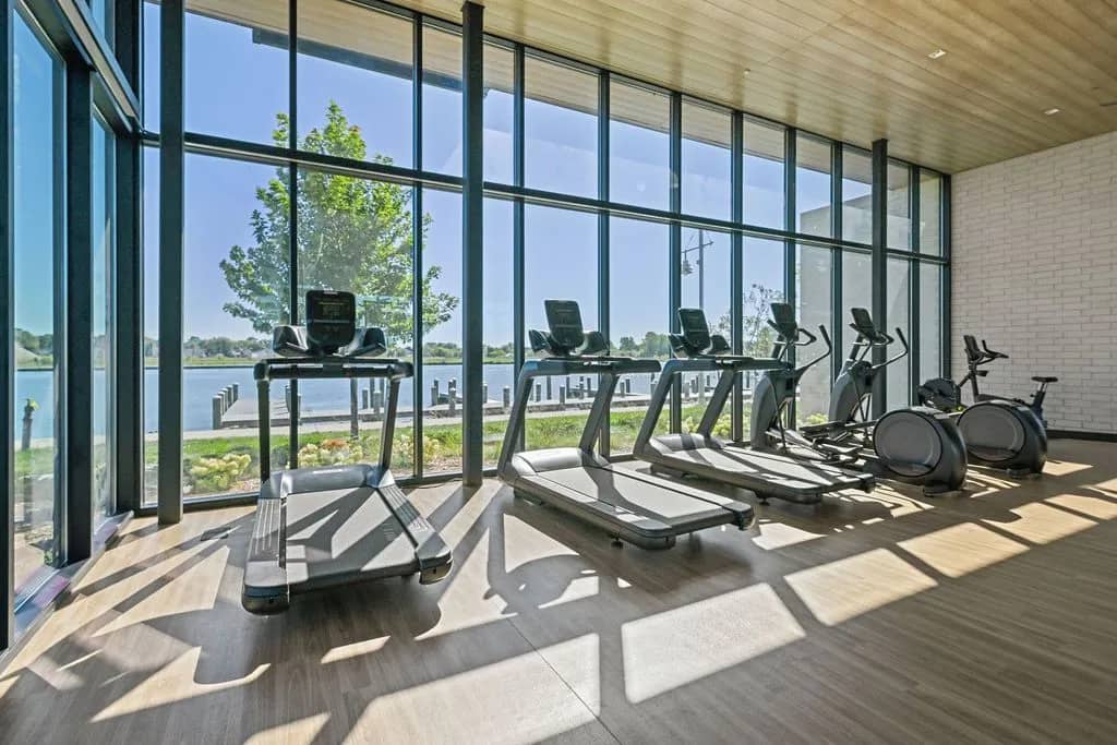 Fitness center with treadmills and exercise bikes facing floor-to-ceiling windows overlooking a lake and green landscape.