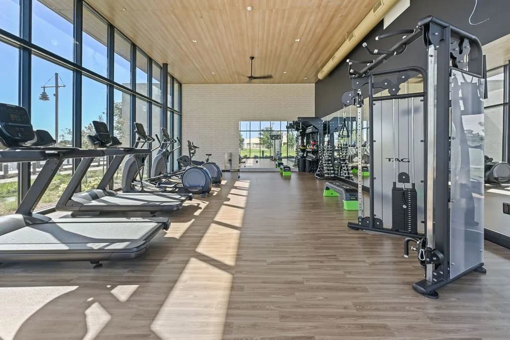 Modern fitness center with multiple treadmills along large windows, weight machines, and wood plank flooring with natural light.