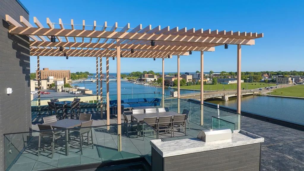 Rooftop terrace with wooden pergola, dining tables, lounge seating, and glass railings overlooking Oshkosh waterfront and river.