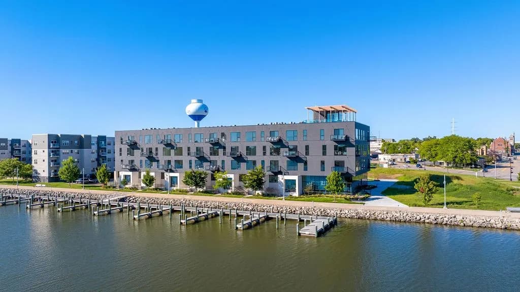 Multi-story apartment building with blue and gray facade overlooking Fox River with wooden boat docks and green landscaping in Oshkosh.