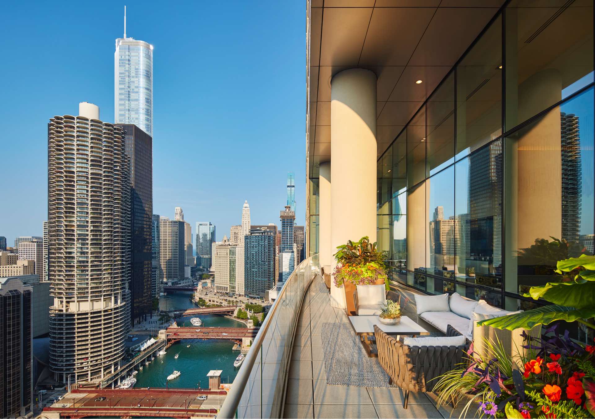 High-rise balcony with seating, potted plants, and flowers overlooking Chicago River and downtown skyline under clear blue sky.