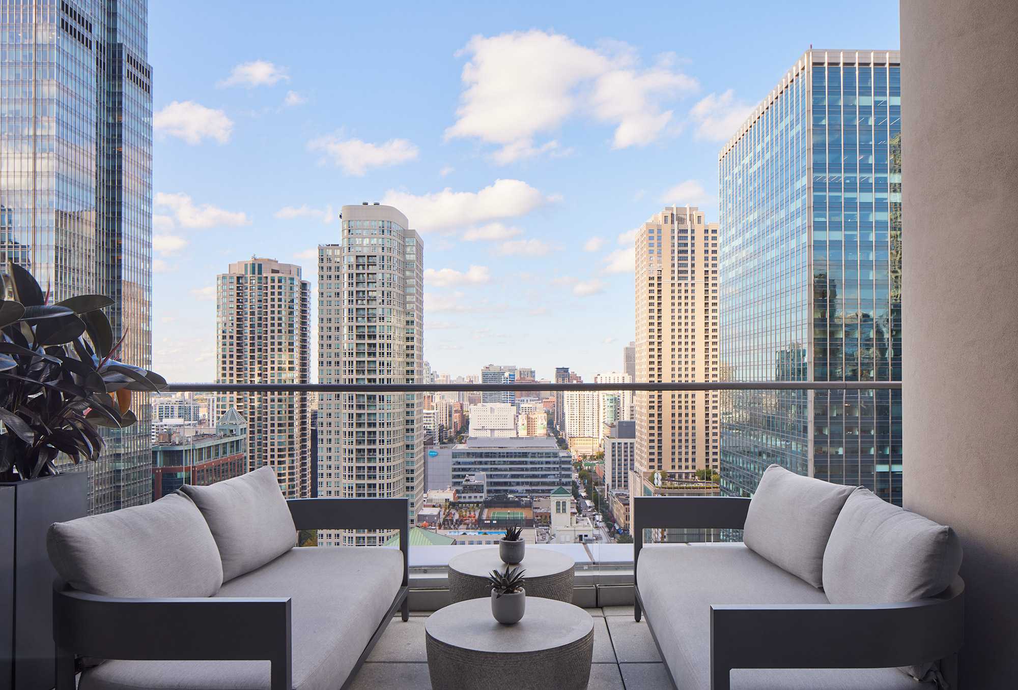 High-rise balcony with two gray upholstered chairs, round table, and expansive view of Chicago downtown skyline and surrounding buildings.