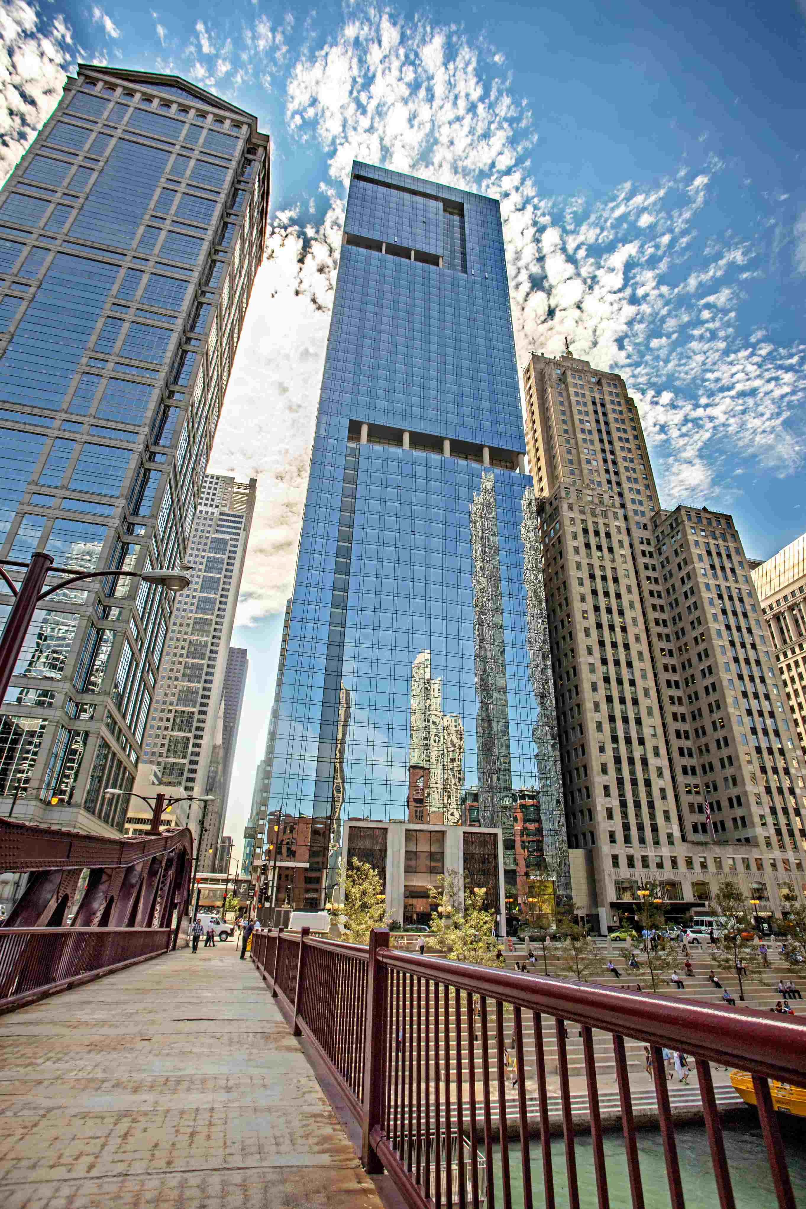 Upward view of modern glass tower with reflected historic building, surrounded by downtown Chicago skyscrapers and pedestrian plaza below.
