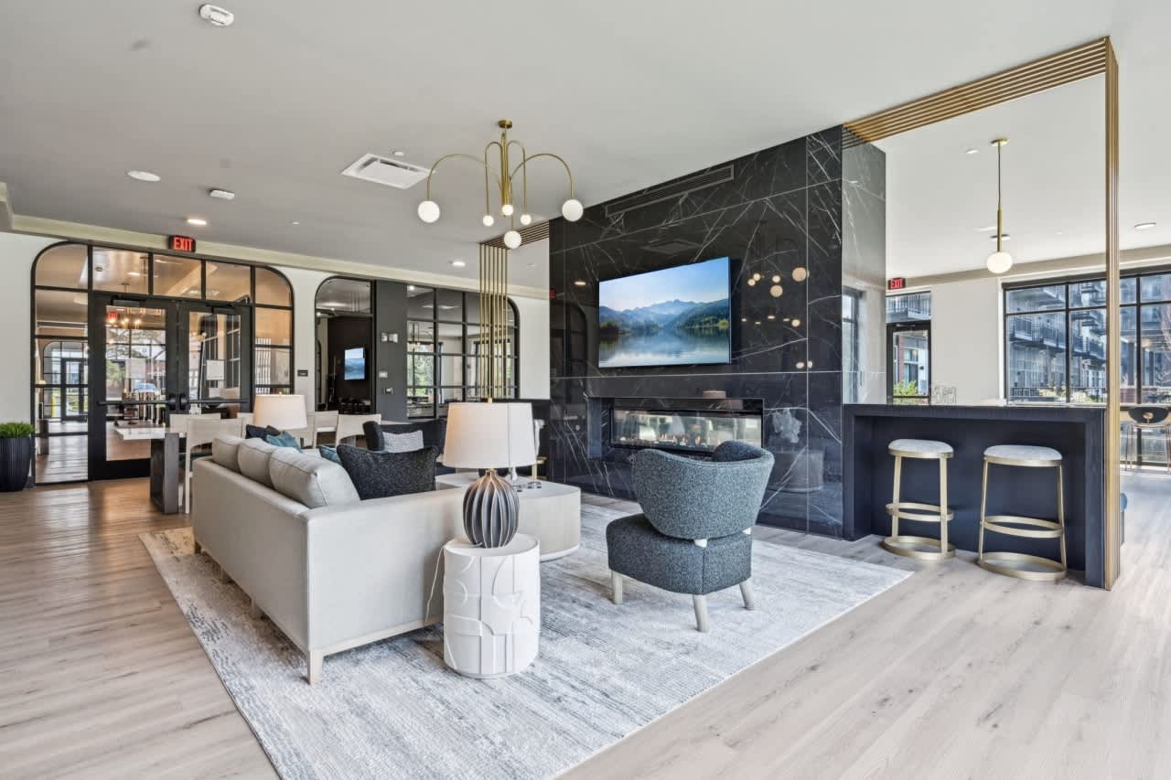 Modern lounge with cream sofa, gray accent chairs, black marble fireplace with TV, and brass pendant lighting overlooking city views.