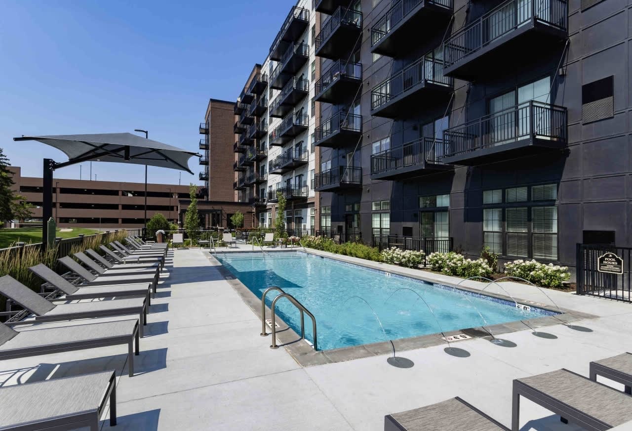 Resort-style swimming pool with lounge chairs, shade structure, and multi-story apartment buildings in background.