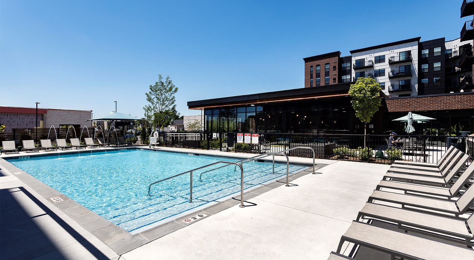 Outdoor swimming pool with metal railings, concrete deck, lounge chairs, and modern building with brick facade in background.