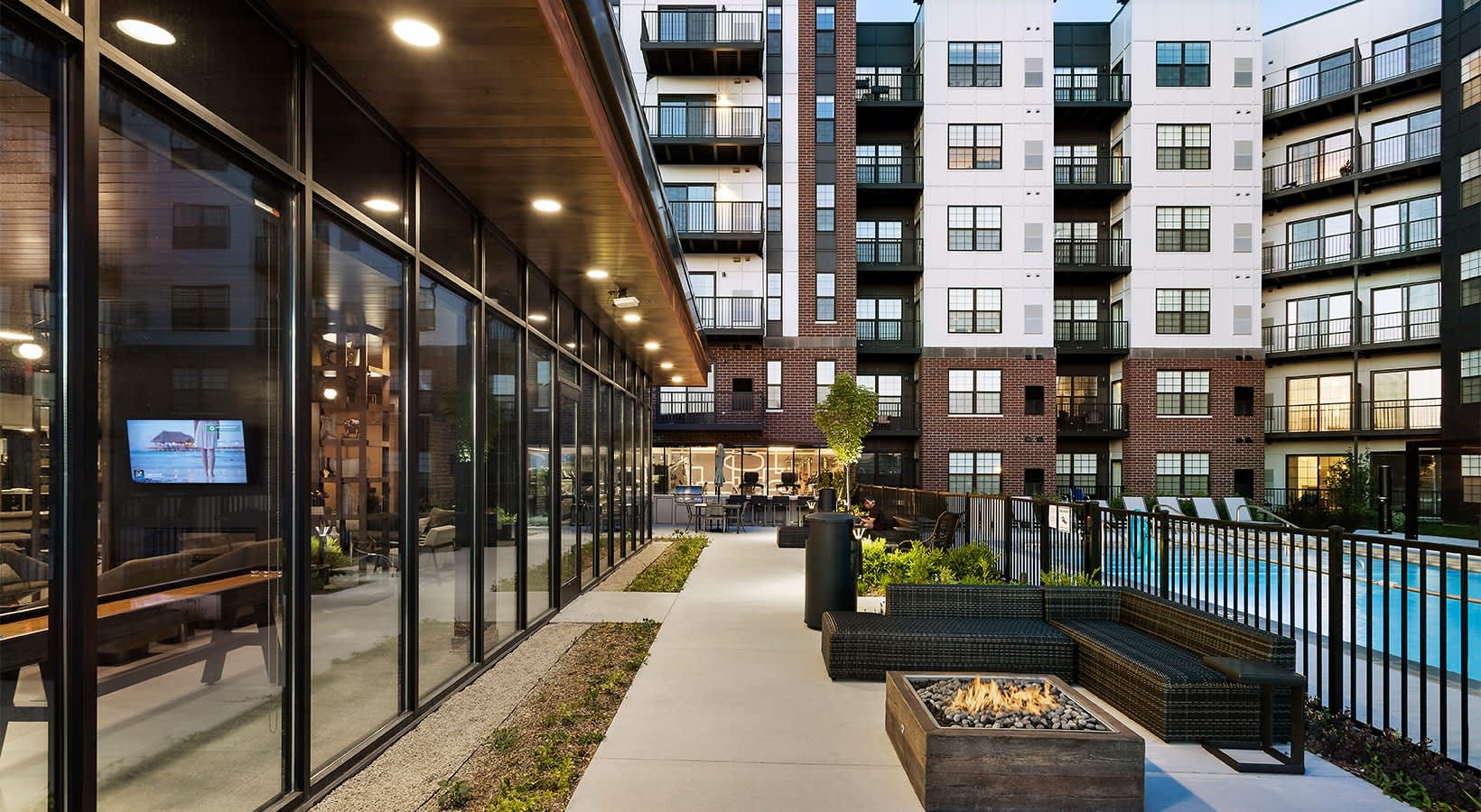 Illuminated courtyard with concrete walkway, fire pit, lounge seating, and pool area bordered by modern residential buildings at dusk.