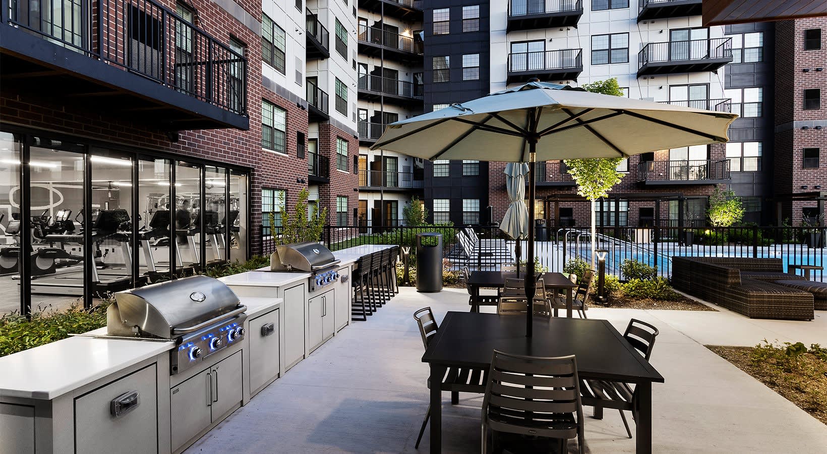 Outdoor courtyard with dining table under umbrella, stainless steel grills, pool, and surrounding residential buildings.