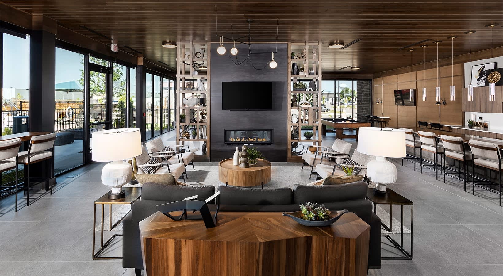 Modern lounge with black sectional sofa, wood coffee table, fireplace with TV, pendant lighting, and glass doors opening to a balcony.