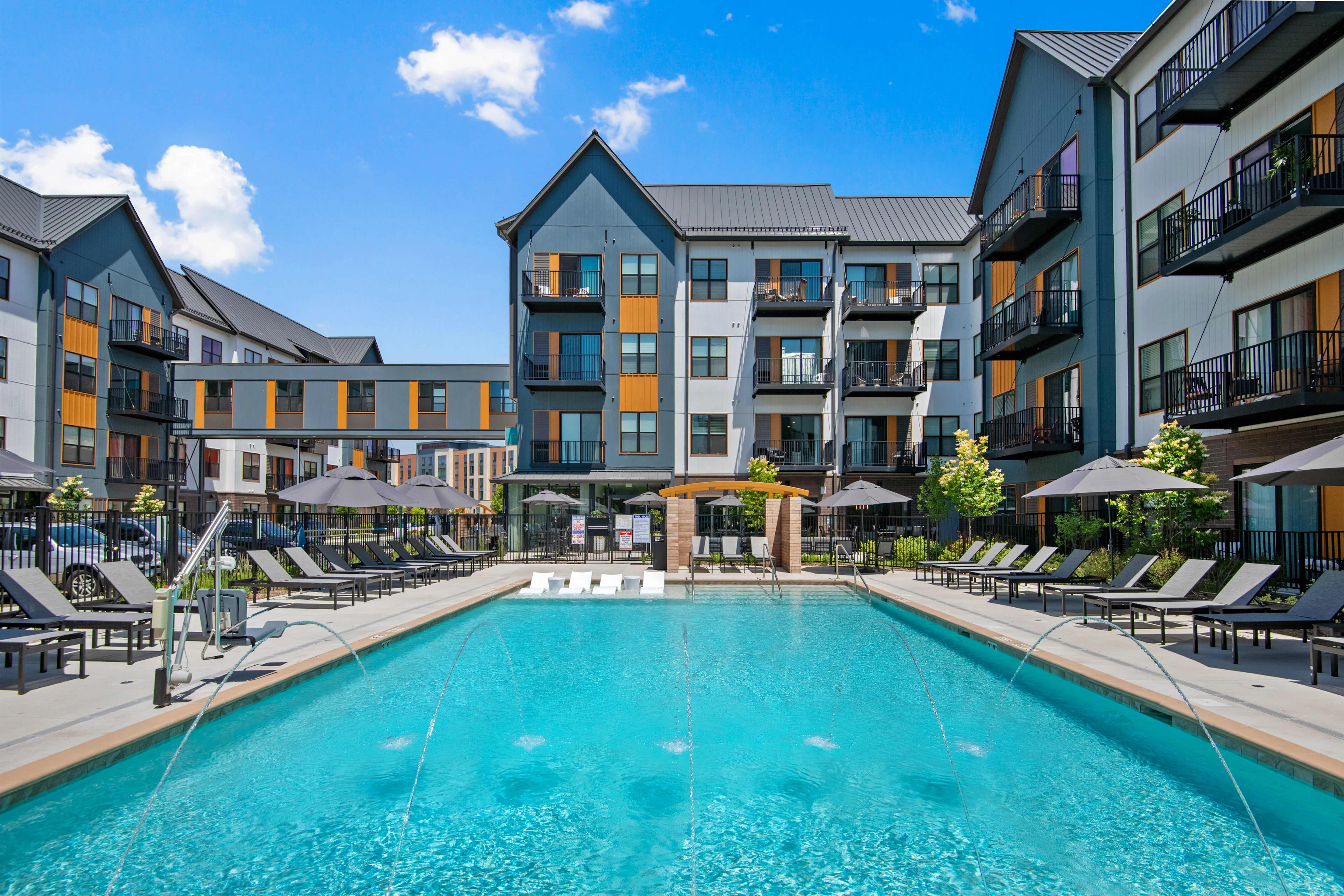 Resort-style swimming pool with clear turquoise water, surrounded by lounge chairs, umbrellas, and multi-story apartment buildings with blue and white facades.