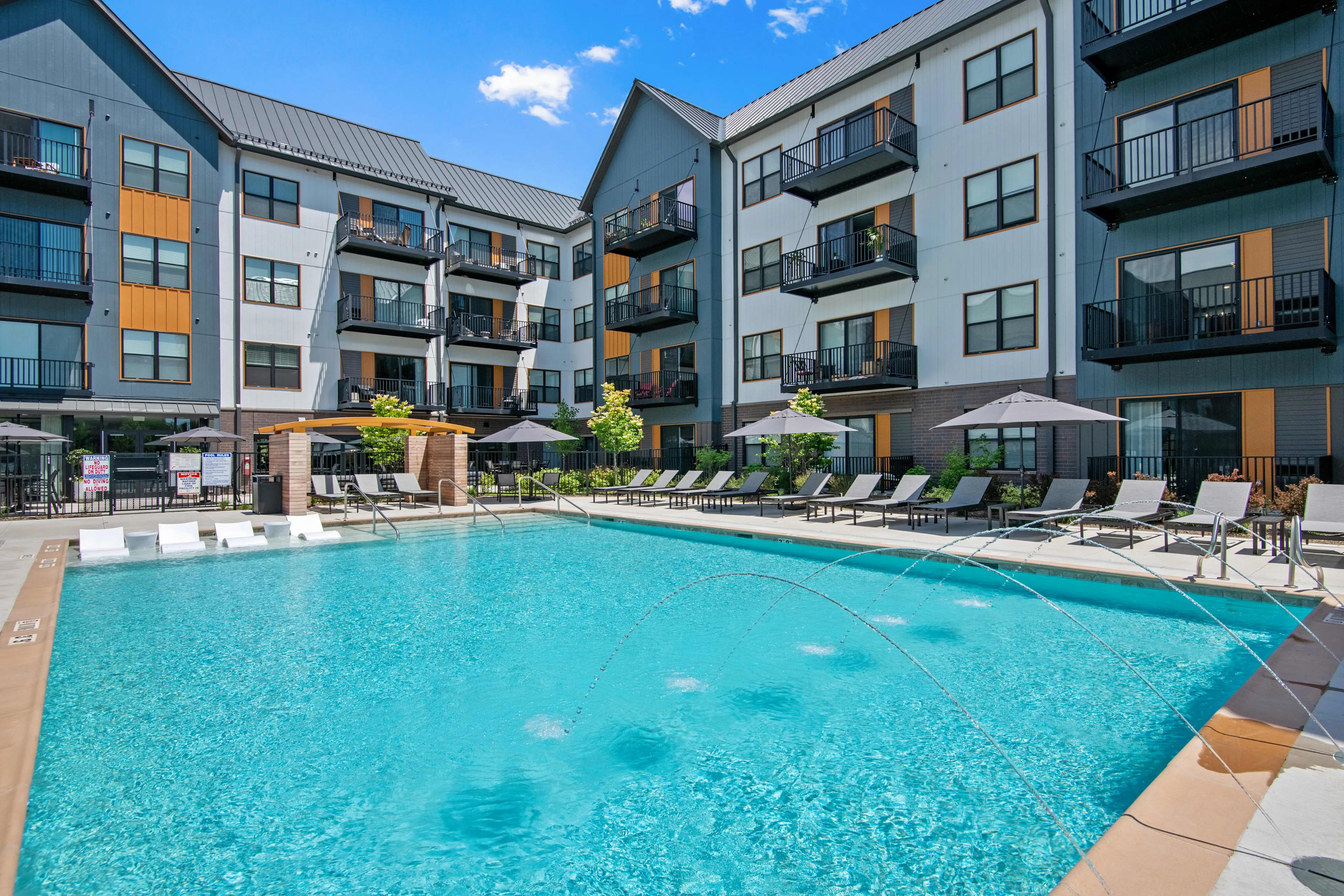 Turquoise swimming pool with lounge chairs and umbrellas, surrounded by multi-story apartment buildings with balconies under clear blue sky.
