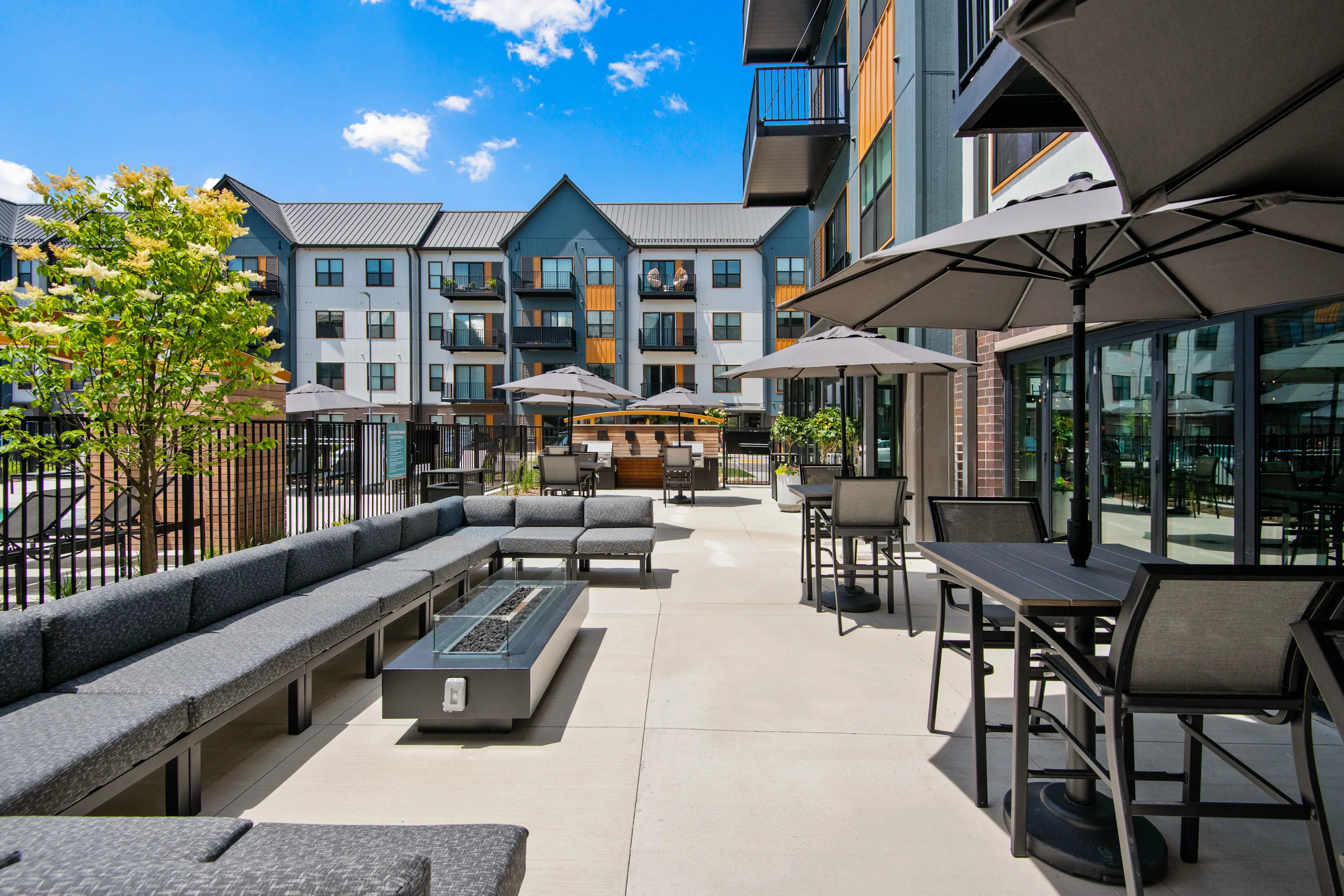 Outdoor courtyard with gray upholstered seating, fire pit, dining tables, umbrellas, and modern apartment building in background under blue sky.