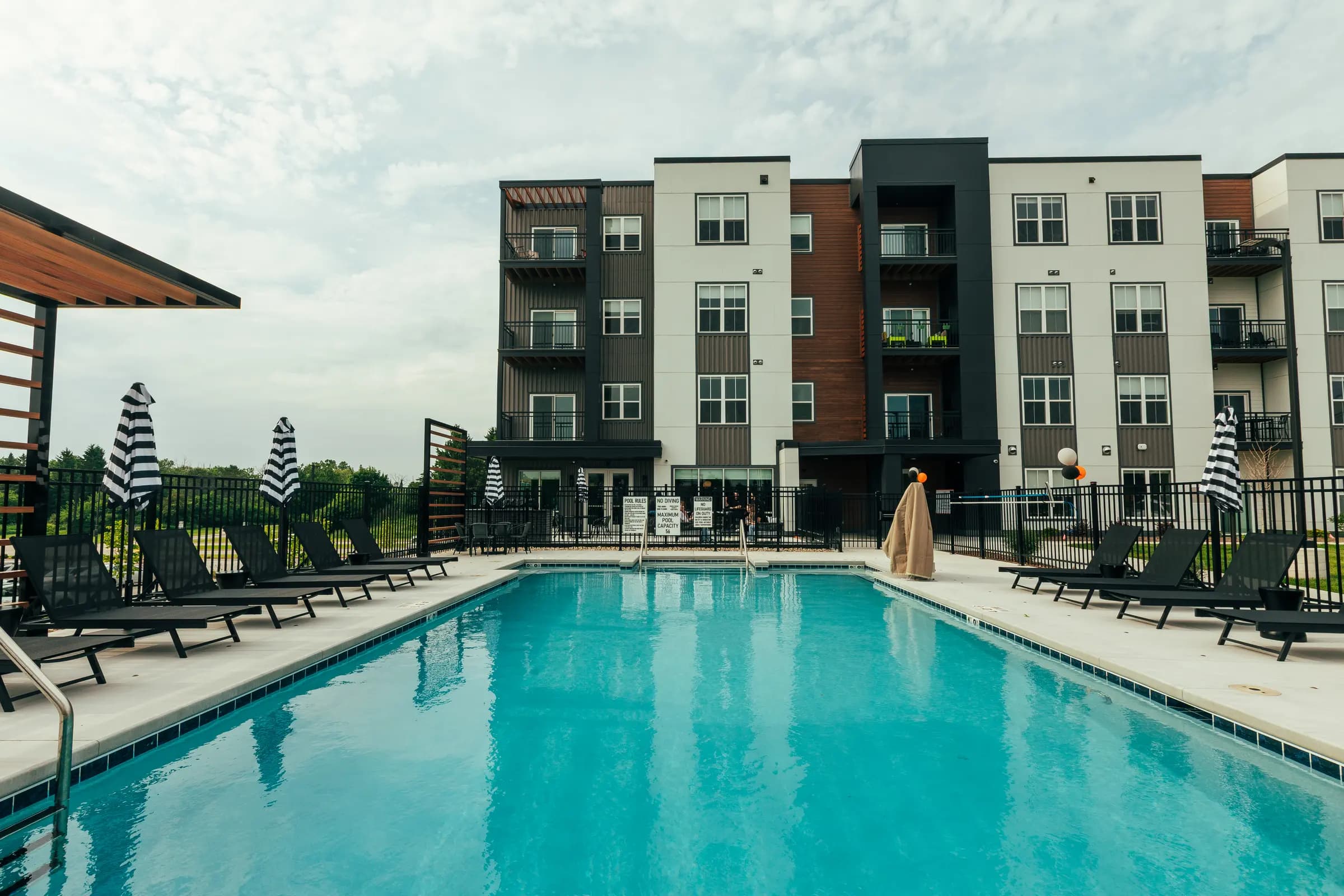 Resort-style swimming pool with turquoise water, black lounge chairs, striped umbrellas, and modern apartment buildings with mixed materials in background.