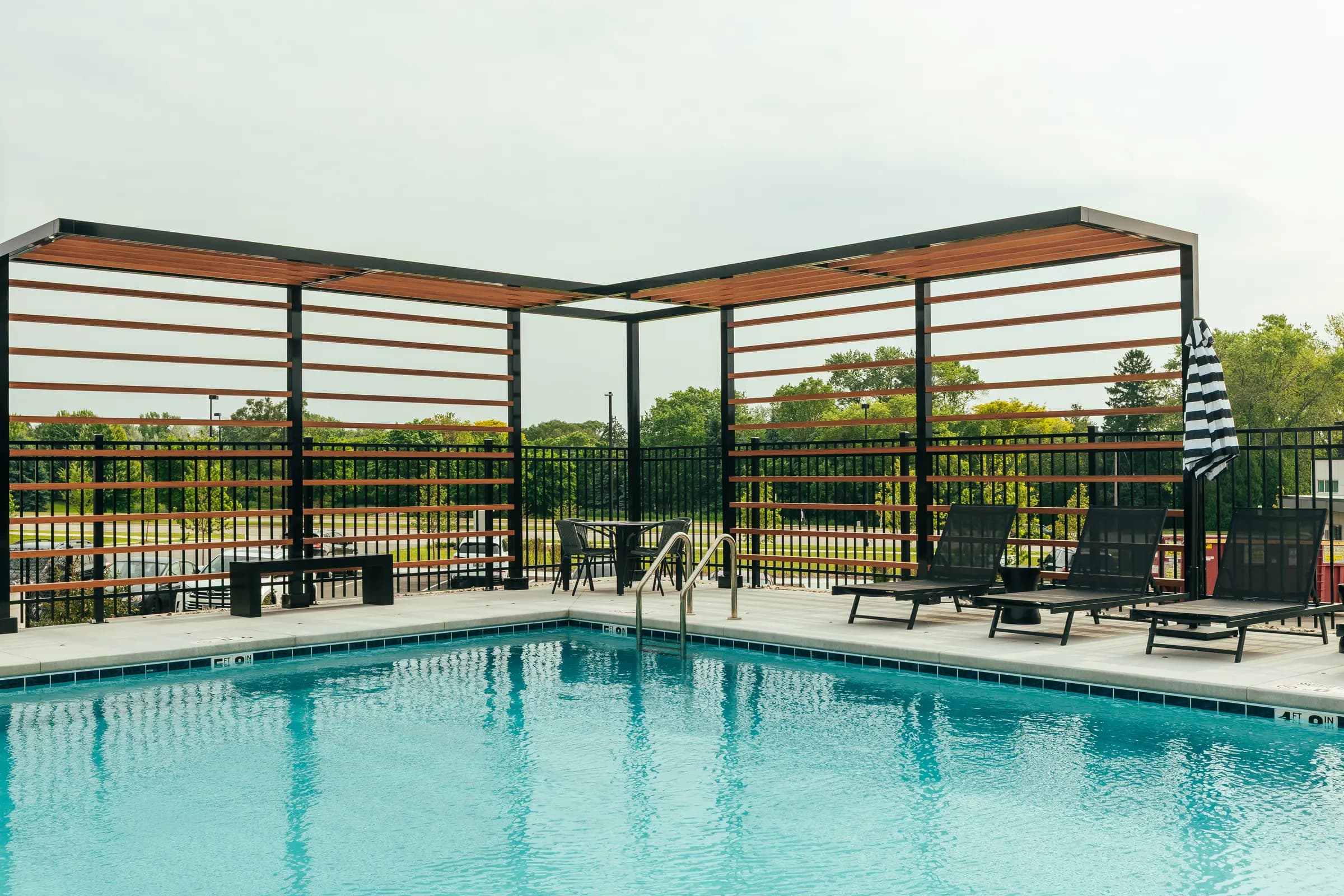 Resort pool with clear turquoise water, black lounge chairs, modern pergola with wood slats and metal frame, metal railing, and green landscape beyond.