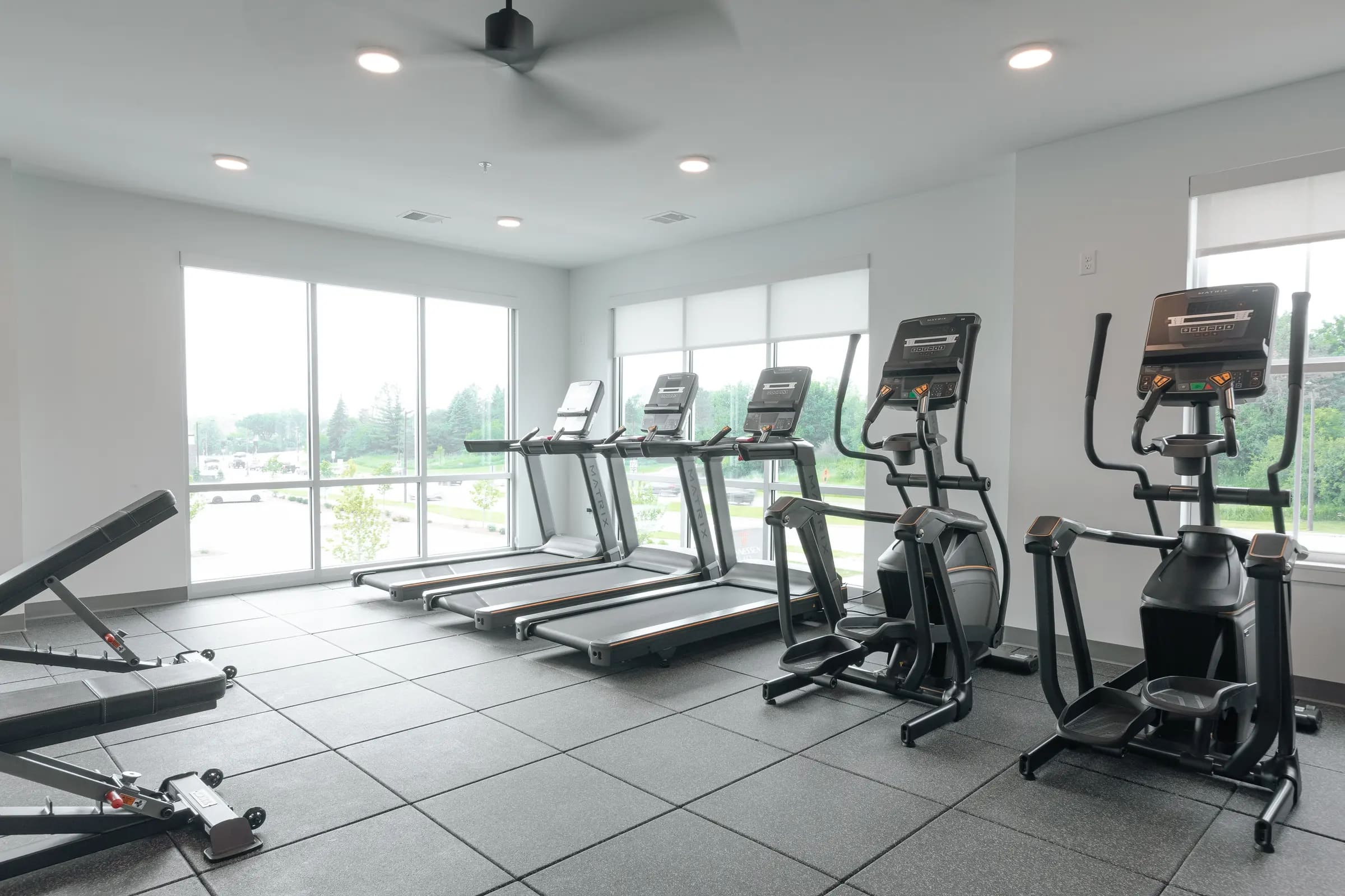 Modern fitness center with multiple treadmills and elliptical machines, large windows, recessed lighting, and tile flooring.