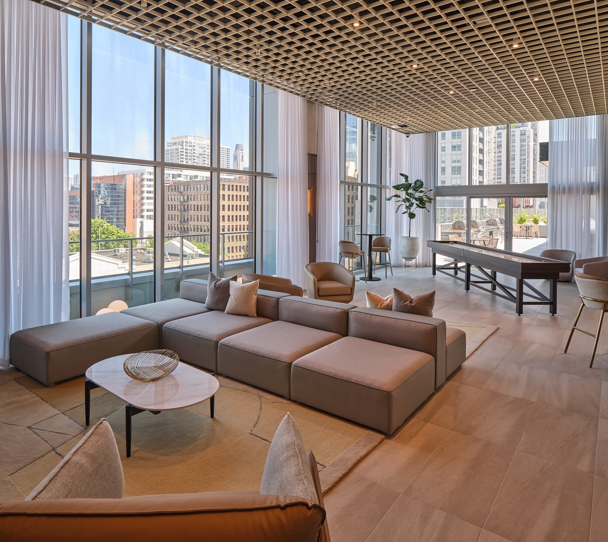 High-rise lounge with brown sectional sofa, wooden coffee table, floor-to-ceiling windows framing Chicago skyline, lattice ceiling, and city views.