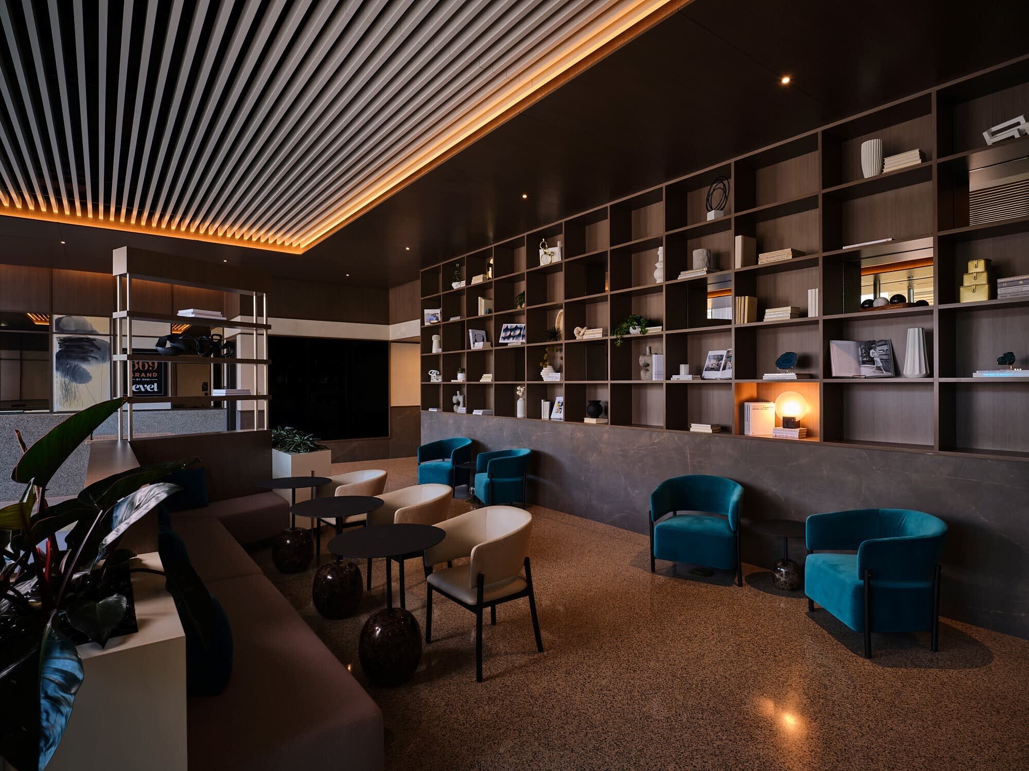 Modern lounge with teal velvet chairs, floor-to-ceiling shelving, linear ceiling lights, and mixed seating areas on cork flooring.