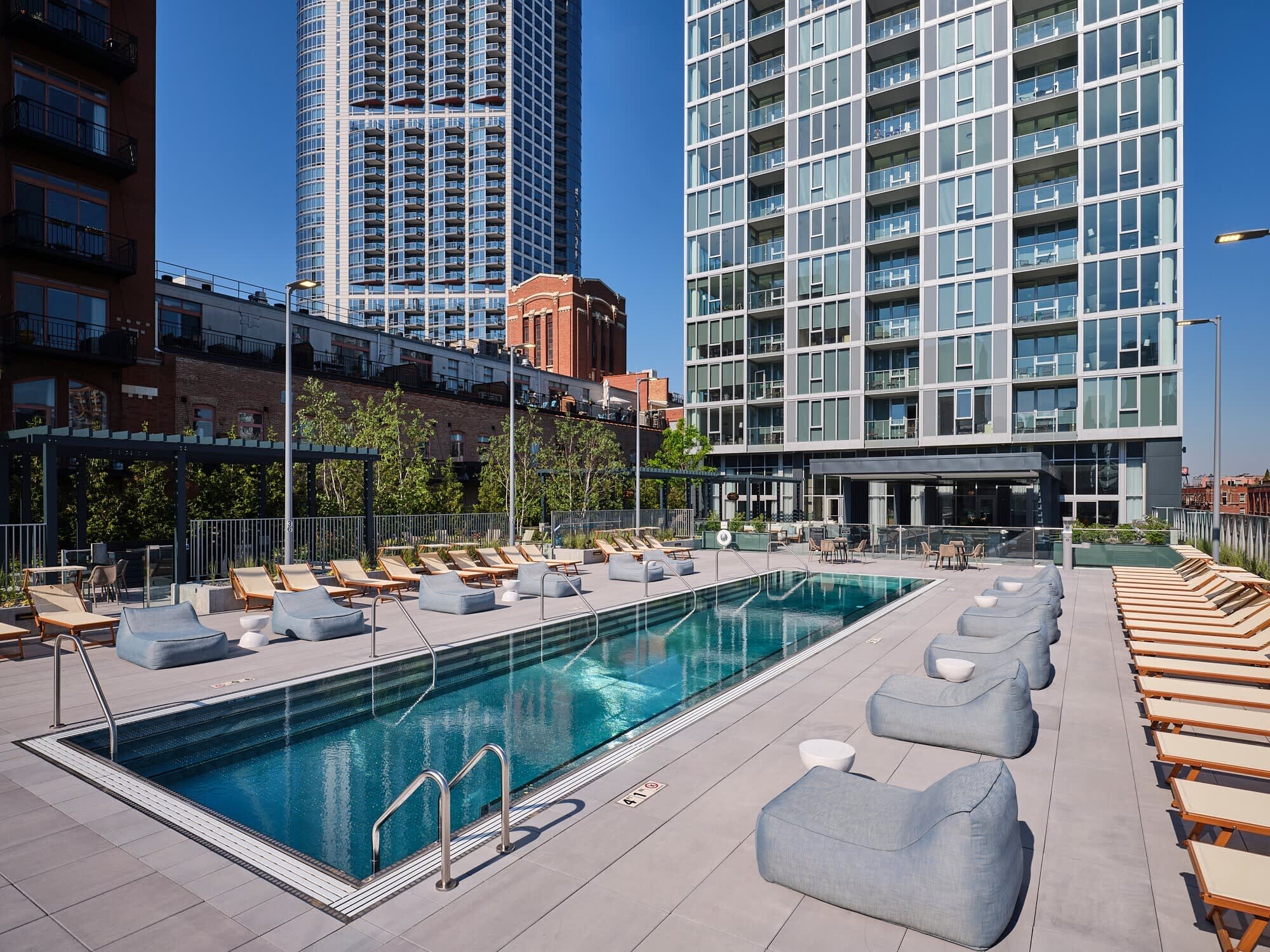 Rooftop pool deck with rectangular swimming pool, gray lounge pods, wooden chaise lounges, and modern high-rise buildings in background under clear blue sky.